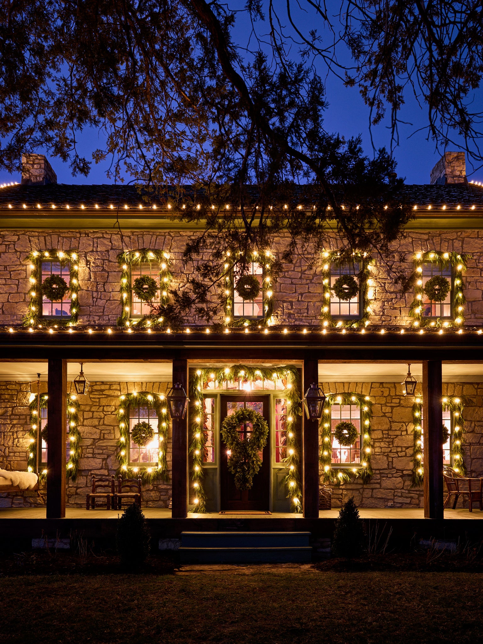 A stone house decorated with glowing white Christmas lights and wreaths on the porch, windows, and door, creating a festive atmosphere at dusk.