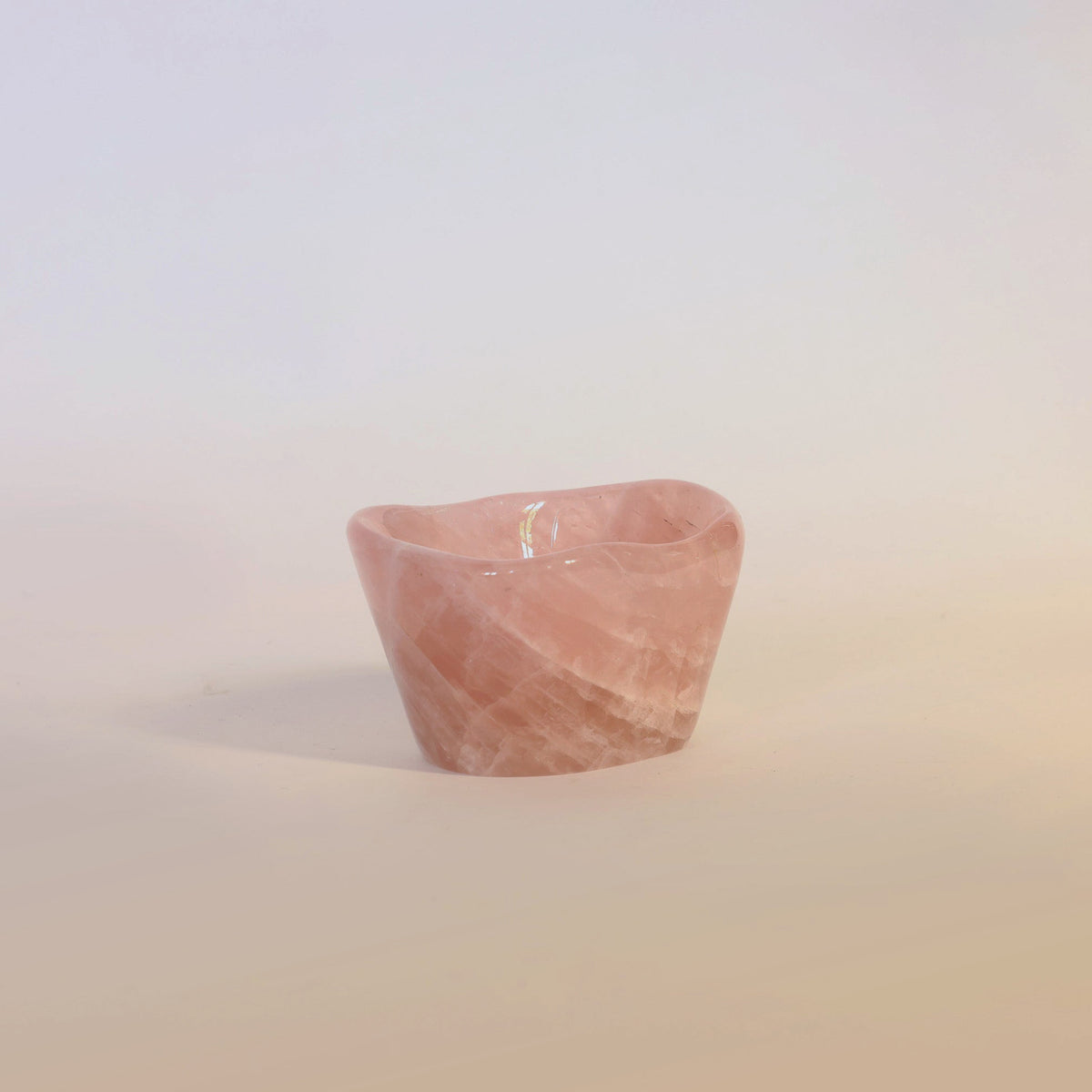 A small, pink, translucent Carved Rose Quartz Bowl by Euro Trash Annie, used as a candle holder with a lit candle inside, sits on a plain light surface casting a soft shadow—a charming decorative accent for any setting.