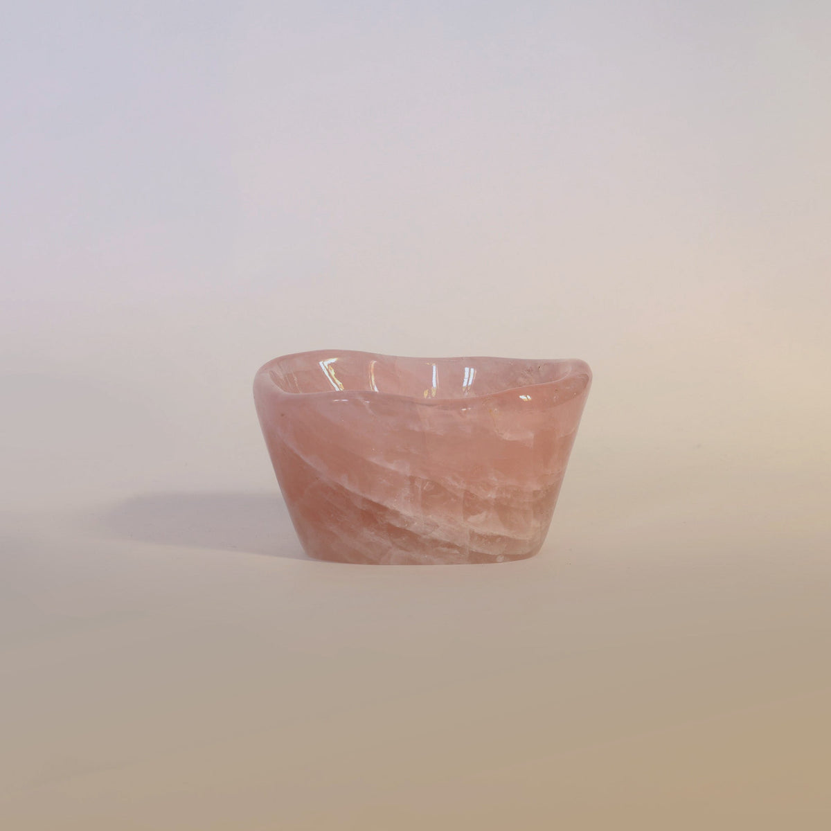 The Carved Rose Quartz Bowl by Euro Trash Annie serves as a decorative accent, sitting gracefully on a light, neutral surface against a soft, gradient background.