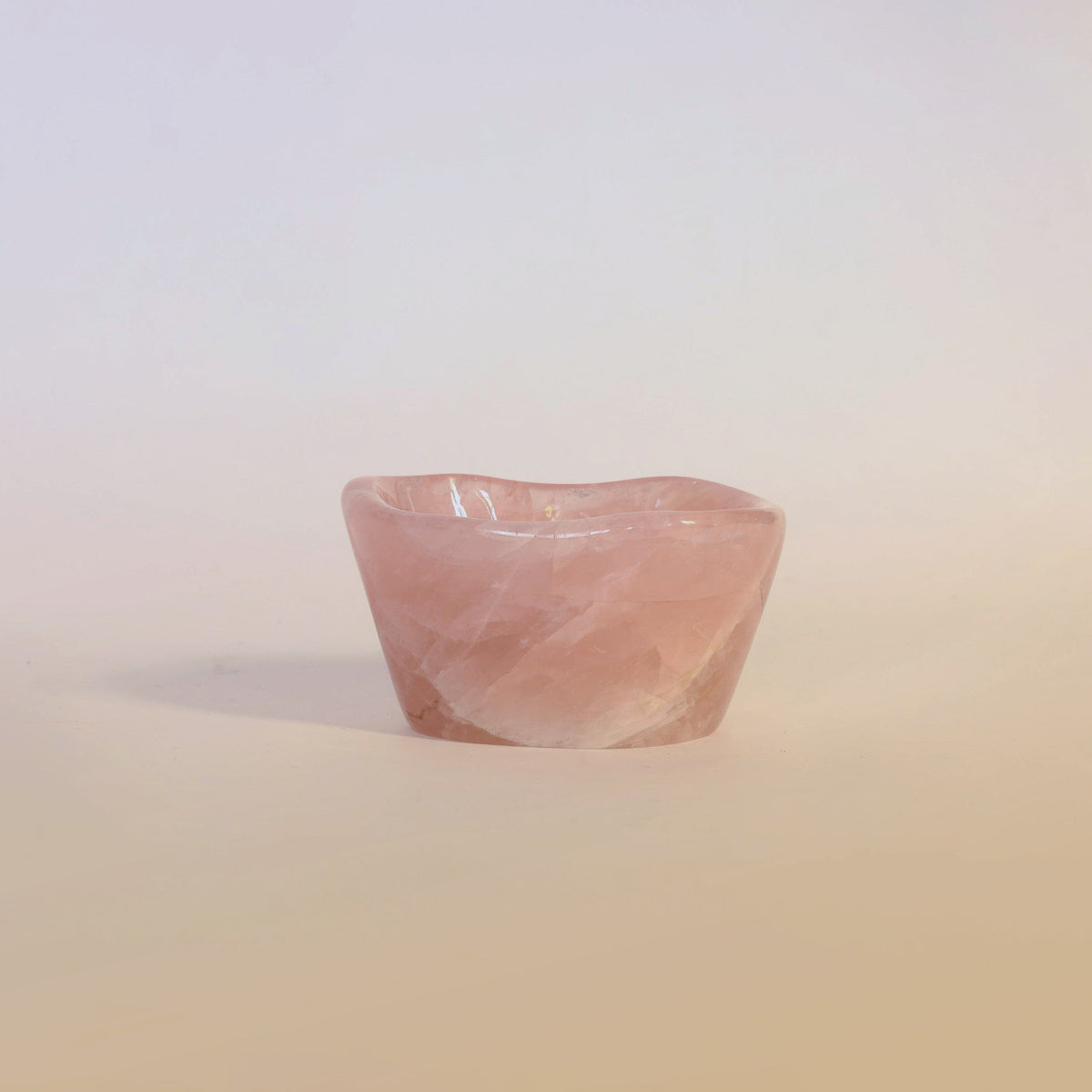 The Carved Rose Quartz Bowl by Euro Trash Annie is a small, polished accent piece, elegantly displayed on a smooth, pale surface with a softly graduated background.