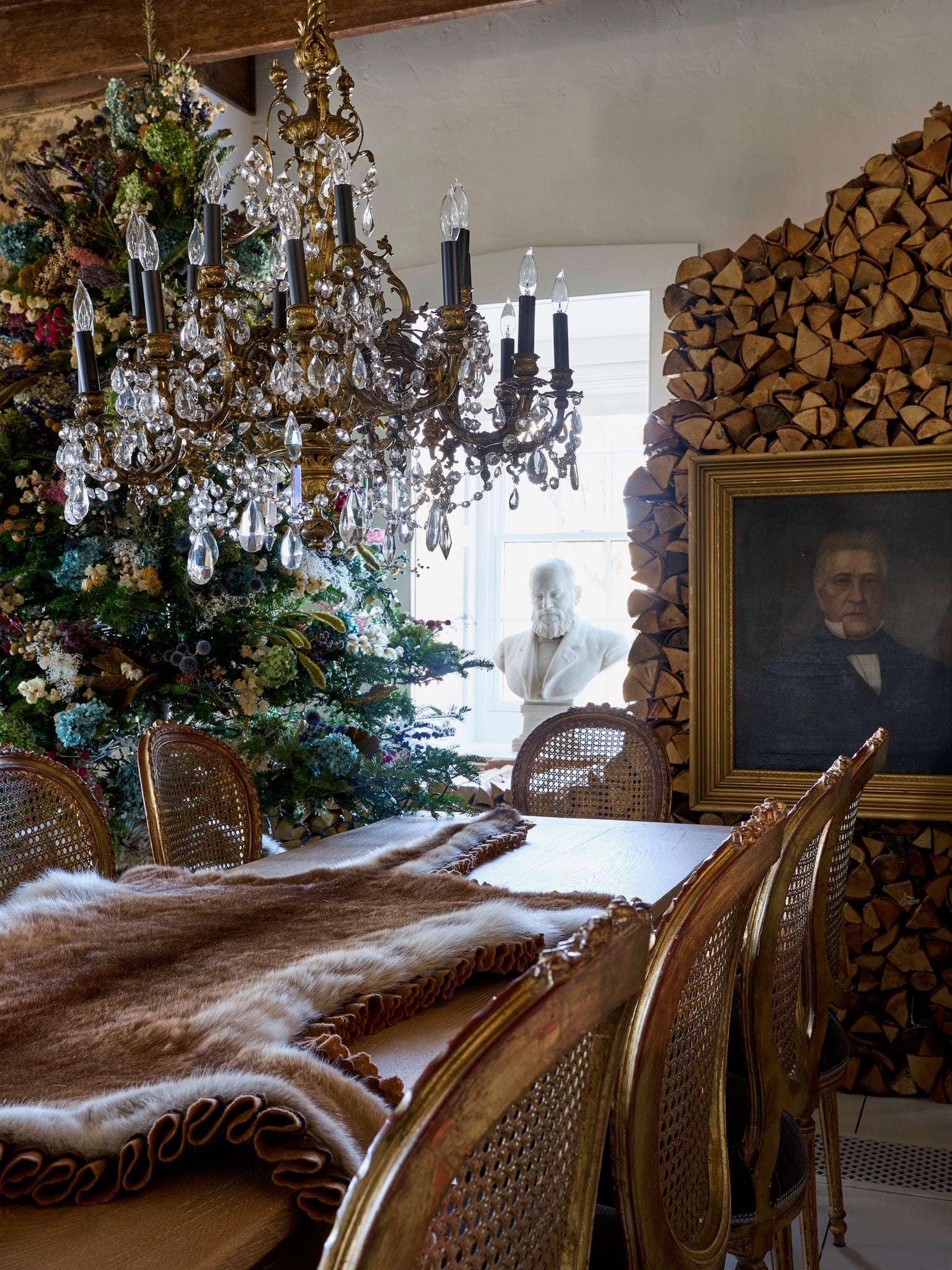 A rustic dining room with a wooden table and cane chairs, a fur throw, a crystal chandelier, stacked firewood, a decorated Christmas tree, a classic portrait, and a white statue by a window in the background.