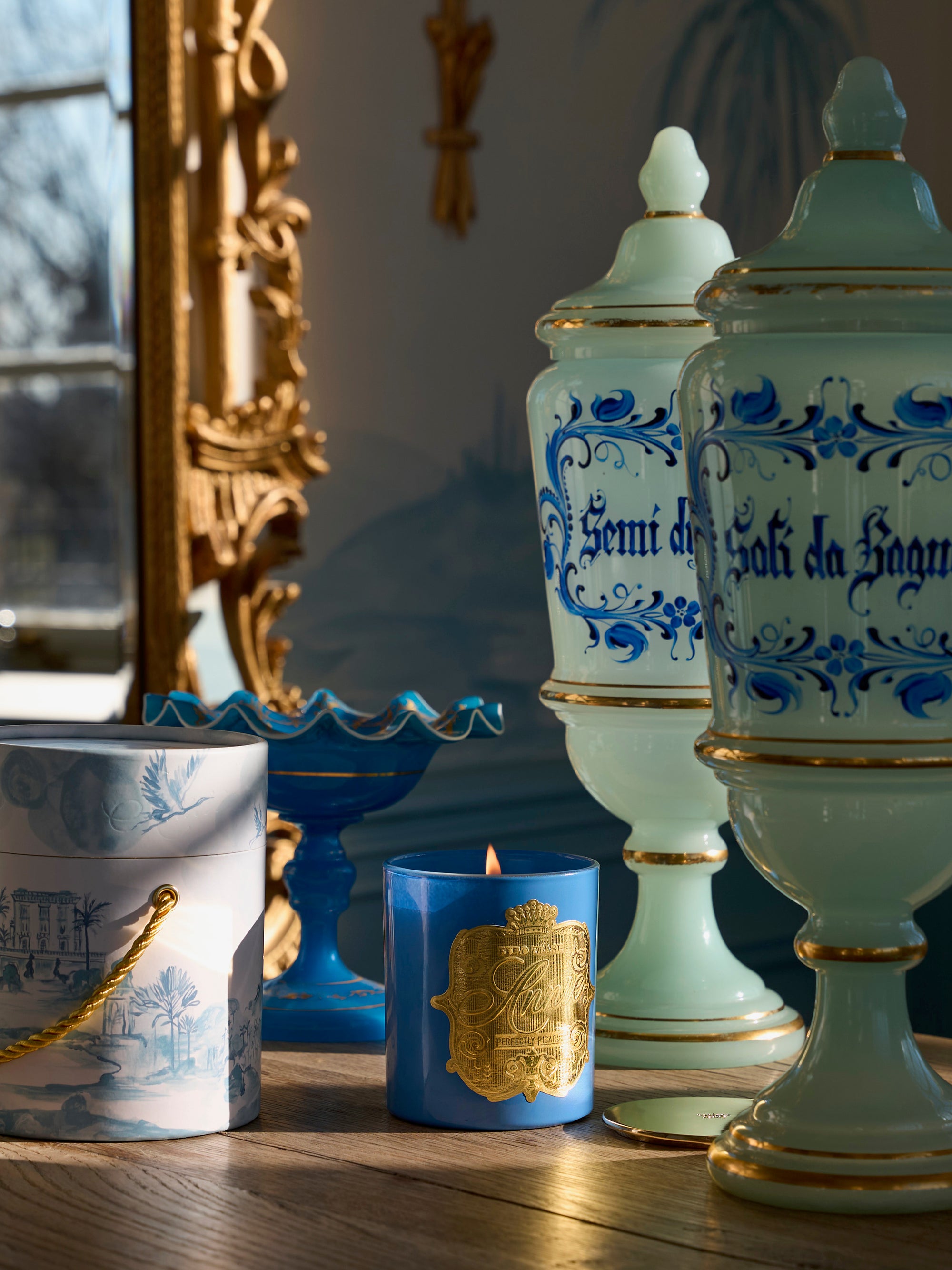 The Perfectly Picardy blue scented soy candle with a gold label burns on a wooden surface, surrounded by ornate pastel jars, a blue dish, and a decorative box as sunlight streams through the window.