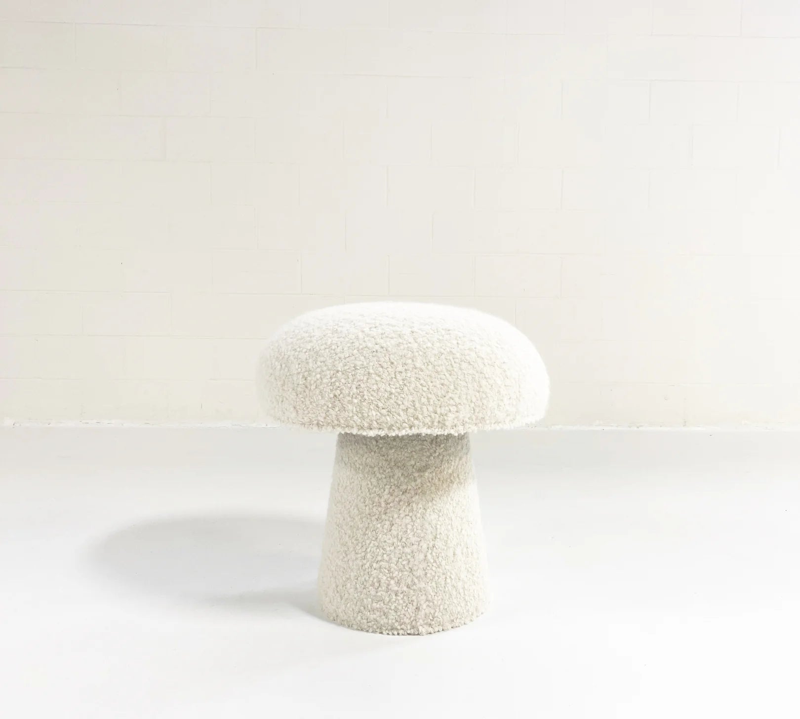 The Forsyth Mushroom Pouf in Boucle, featuring a white textured finish with a rounded top and conical base, sits on a white floor against a plain white wall.