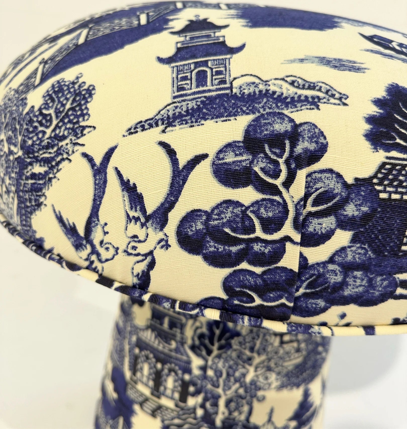 A close-up of The Forsyth Mushroom Pouf in Beata Heuman Willow Linen Cotton, featuring a blue and white pastoral toile scene with trees, pagoda, and foliage in chinoiserie style.