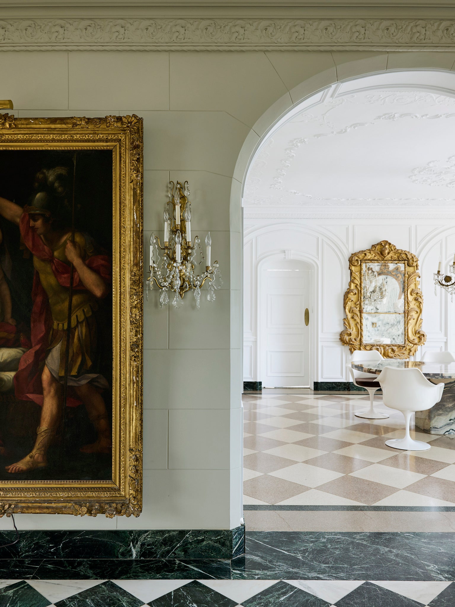 A grand hallway with marble floors, an ornate gold-framed painting and wall sconce on the left, and an arched doorway leading to a bright room with white walls, a large gold-framed mirror, and white modern chairs.