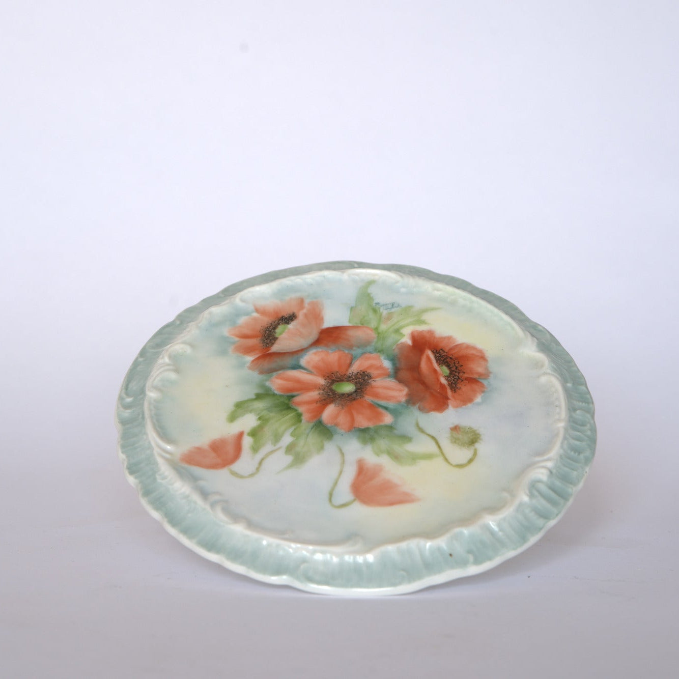 The Euro Trash Vintage Hand-Painted Floral Ceramic Plate features a scalloped light blue rim and a central design of three red-orange poppy flowers with green leaves on a pale background.