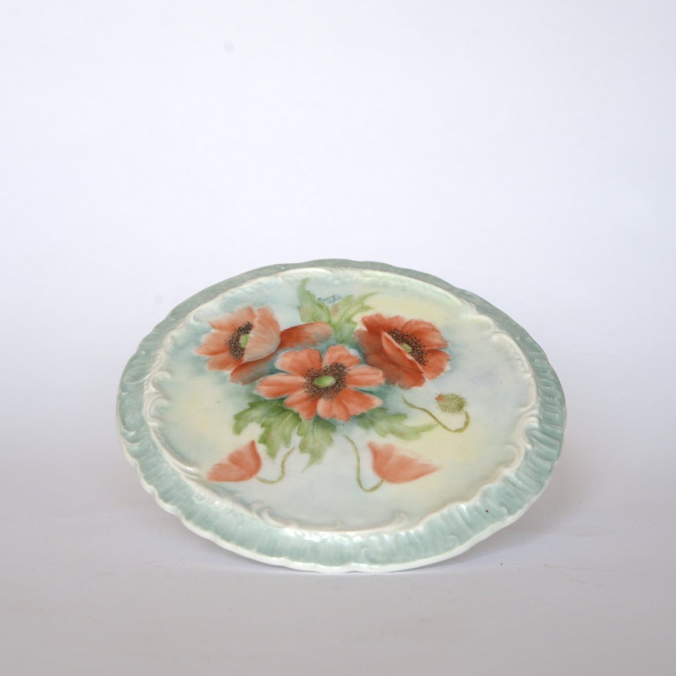 The Euro Trash Vintage Hand-Painted Floral Ceramic Plate features a scalloped edge and a design of red-orange poppies with green leaves on a pale blue and white background.