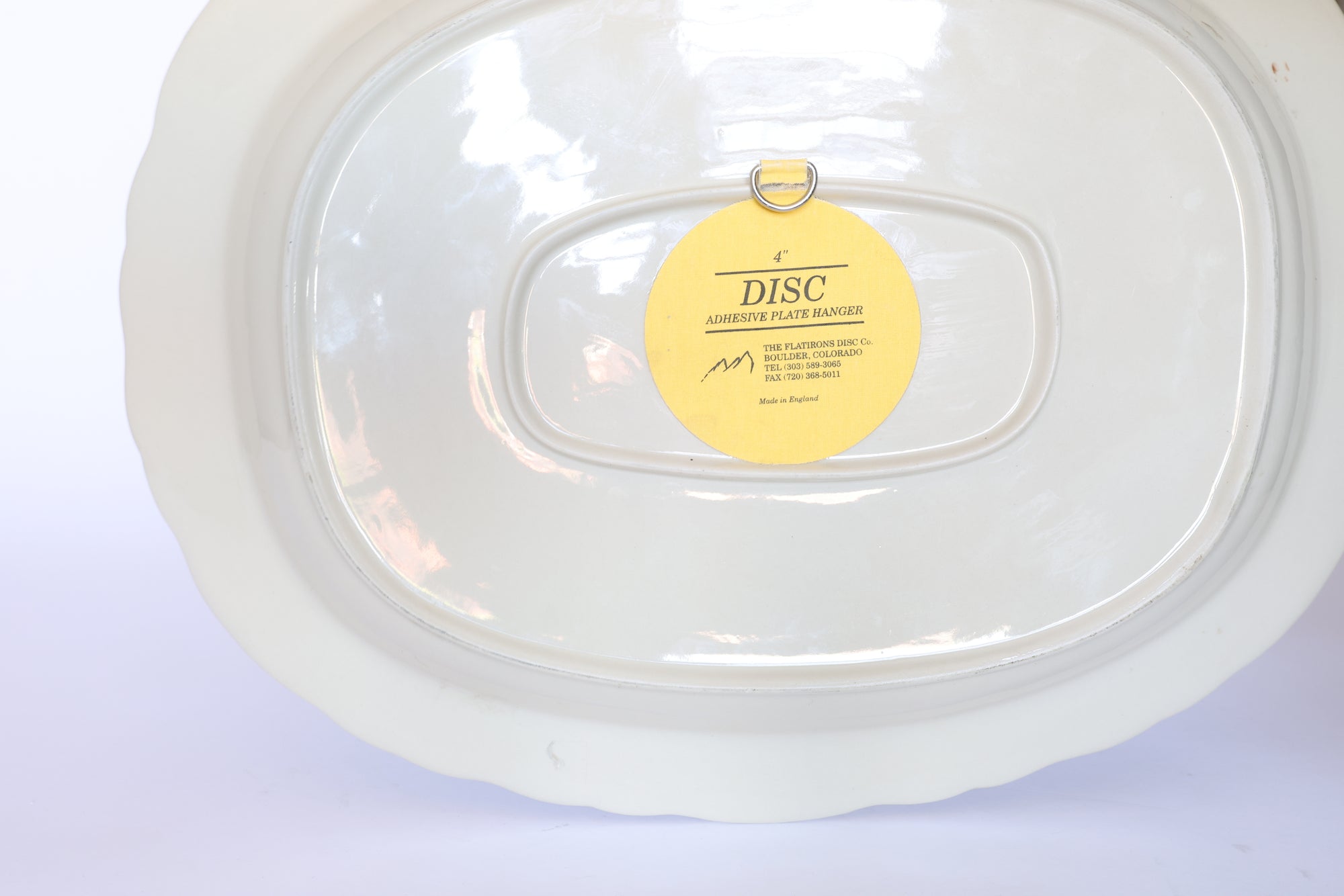 The underside of the Vintage Blue Transferware Oval Serving Platter with Pastoral Scene by Euro Trash features a yellow "DISC ADHESIVE PLATE HANGER" sticker and a central metal loop for hanging.
