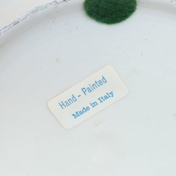 The Vintage White & Blue Ribbon Ceramic Cachepot by Euro Trash features a white ceramic surface with a "Hand-Painted, Made in Italy" blue sticker and a green felt pad partially visible at the top, evoking classic vintage style.