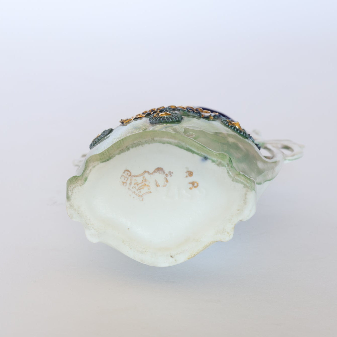 A close-up of the underside of a Vintage Blue & Gold Porcelain Trinket Dish by Euro Trash, featuring detailed blue and gold accents on the sides and a faded maker's mark on the white base. The background is plain and light colored.