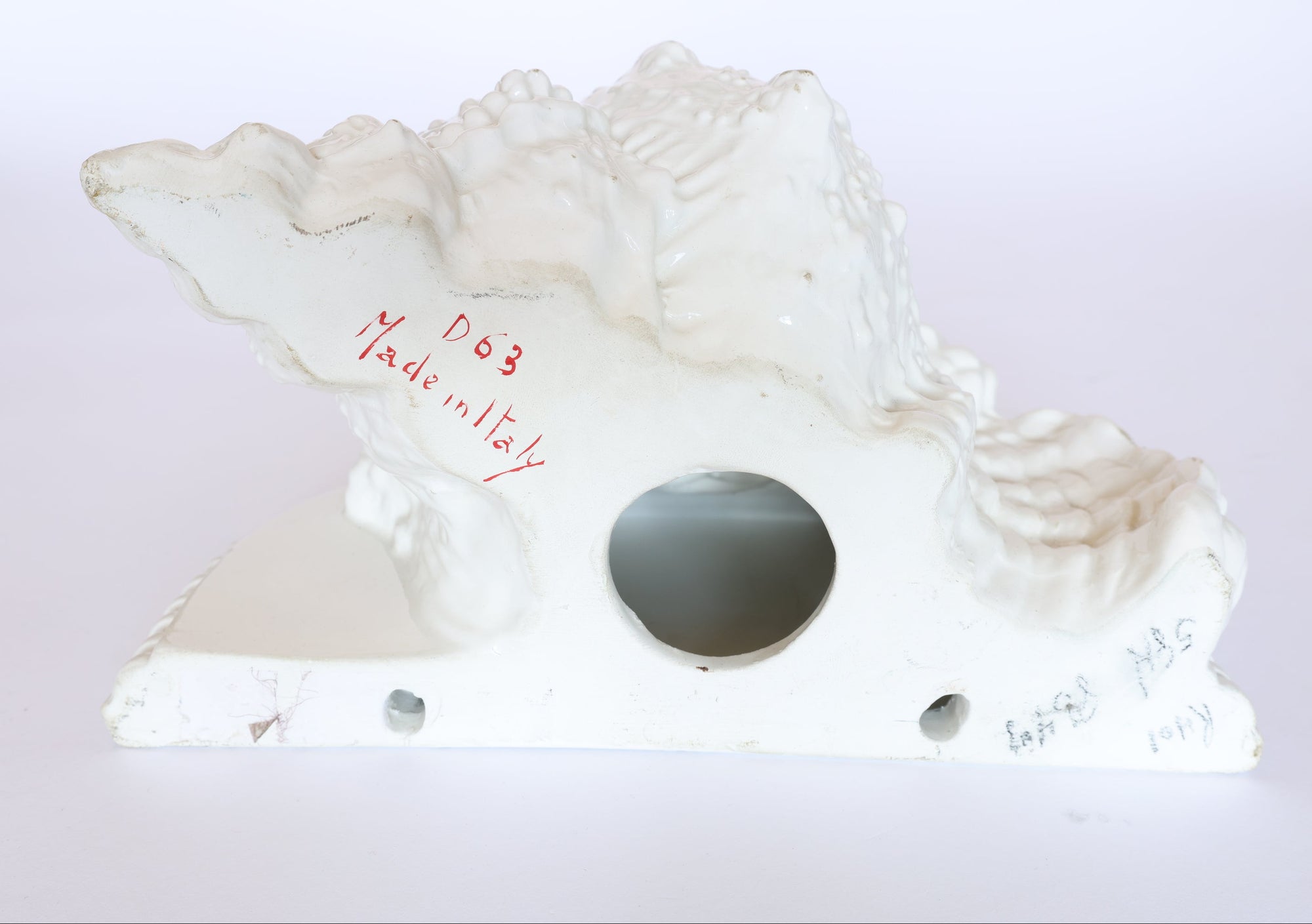 The underside of this Vintage Milk Glass Seashell Sculpture by Euro Trash Annie shows "D63 Made in Italy" handwritten in red, faint pencil marks, a central round hole on the base, and two smaller edge holes—perfect as a coastal accent.