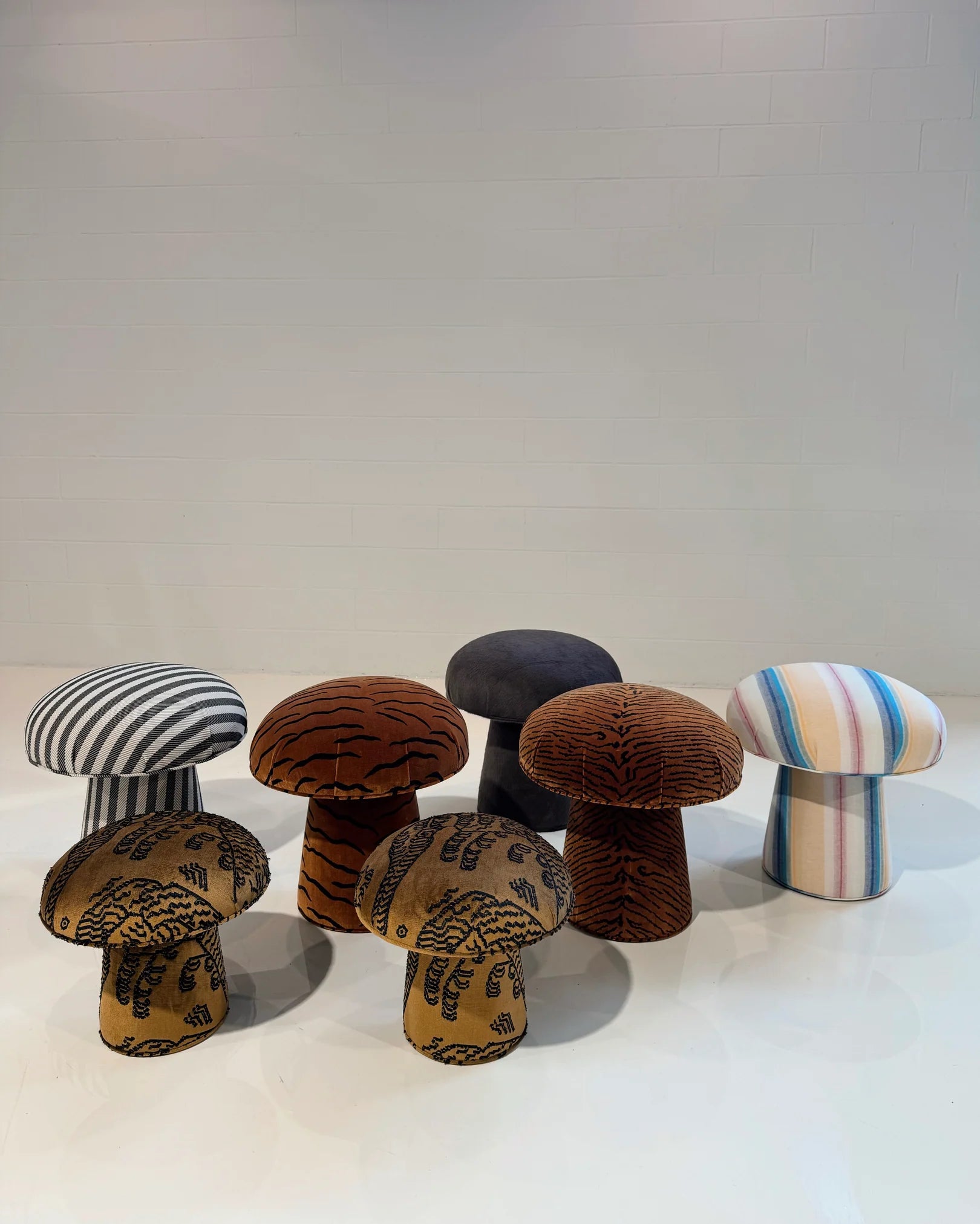 Six mushroom-shaped stools, including a handmade ottoman and The Forsyth Mushroom Pouf in Dedar 'A Tiger In The Orangerie' jacquard velvet, with striped and abstract patterns, are arranged on a shiny white floor against a plain white wall.