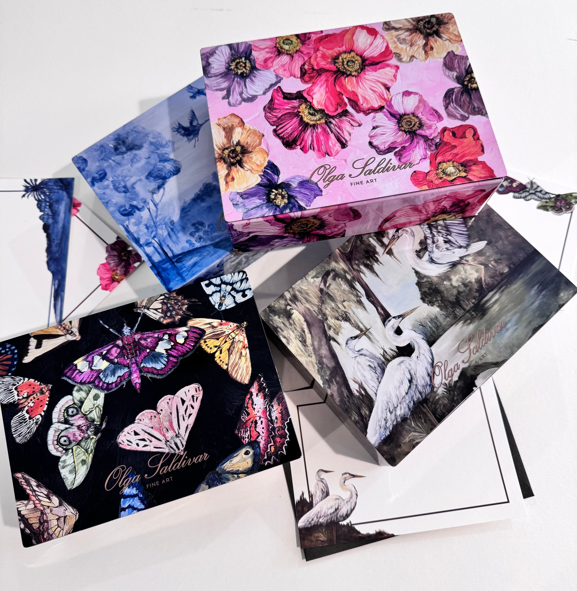 Three decorative boxes from the Euro Trash Annie Poppy Serenade Stationery Set—adorned with pink flowers, butterflies, and white birds—sit atop matching art prints and floral notecards, all elegantly displayed on a white surface.