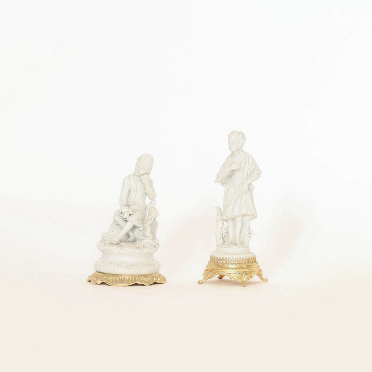 Elevate your collectible home decor with this Pair of Antique Bisque Porcelain Figurines on Gilt Bases by Euro Trash Annie. One figurine features a seated figure with a lamb, while the other portrays a standing figure holding an object, also accompanied by a lamb at their feet. Both are crafted in elegant white bisque porcelain with gilt bases and displayed against a plain white background, making them exquisite additions to any collection.