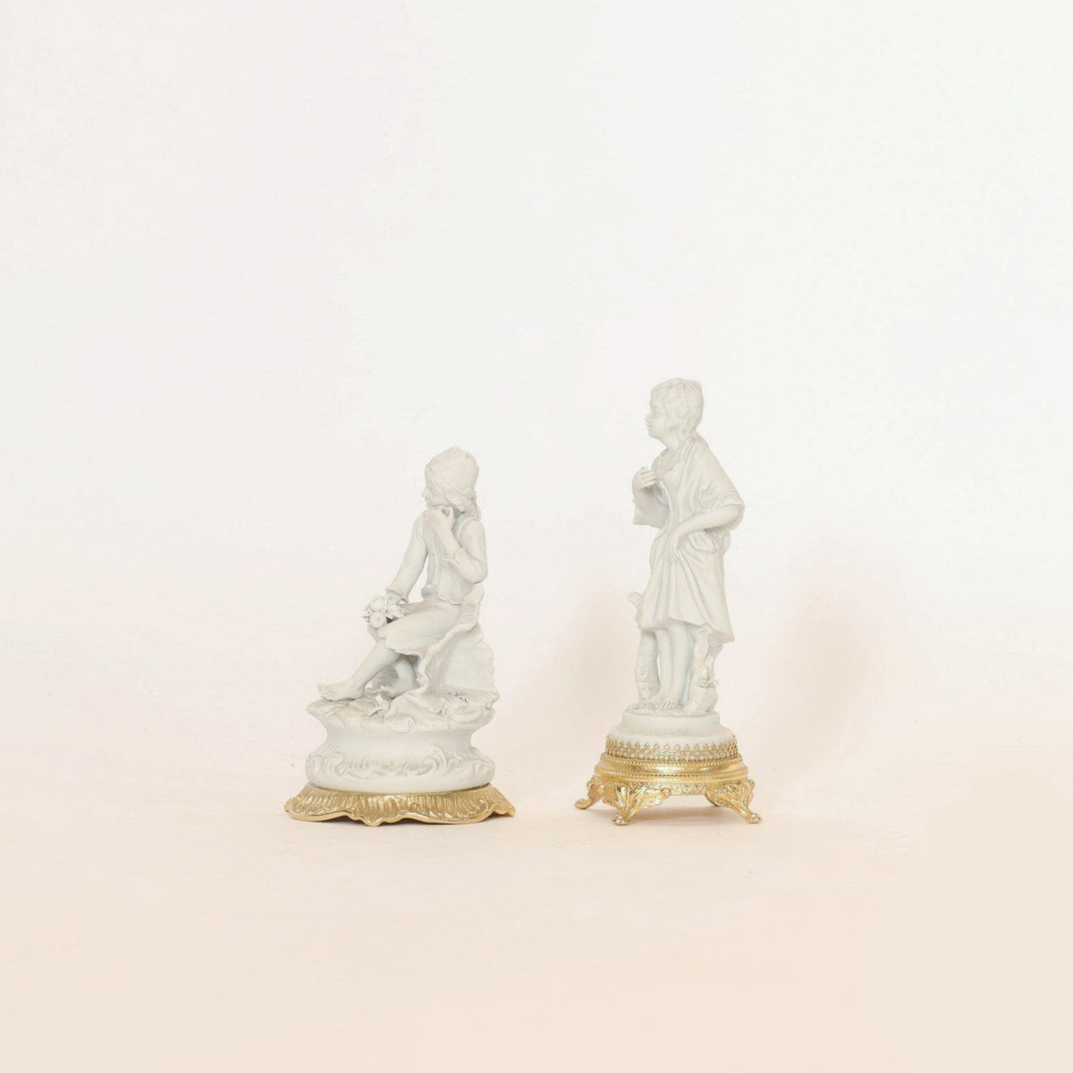 The Pair of Antique Bisque Porcelain Figurines on Gilt Bases by Euro Trash Annie features two collectible home decor pieces: one showcases a seated figure holding flowers, while the other depicts a standing figure with an object. Both are displayed against a plain white background.