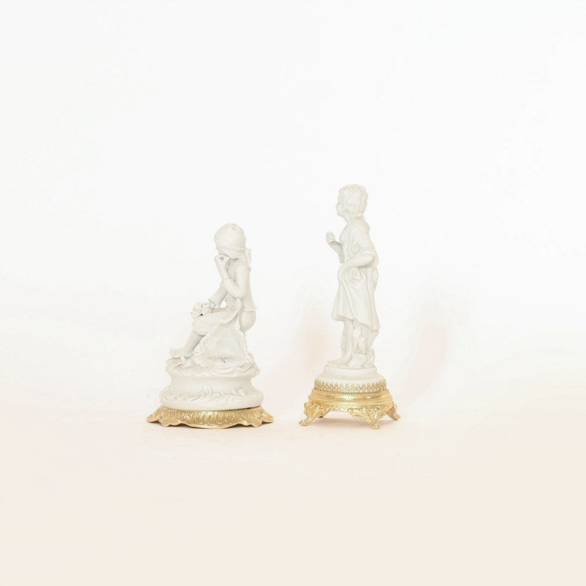 A pair of antique bisque porcelain figurines on gilt bases by Euro Trash Annie features a seated woman holding flowers and a standing man, both ornately crafted and presented against a simple, light background.