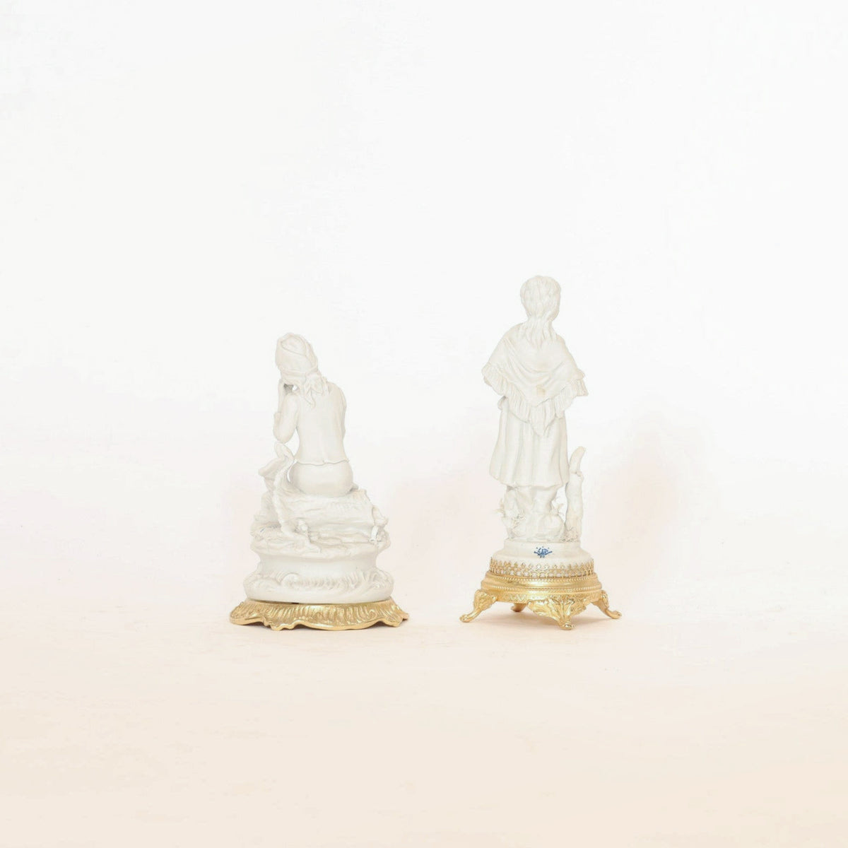A pair of antique bisque porcelain figurines on gilt bases by Euro Trash Annie, displayed side by side against a plain white background. One figurine kneels while the other stands, both facing away—charming and distinctive pieces ideal for collectible home decor.