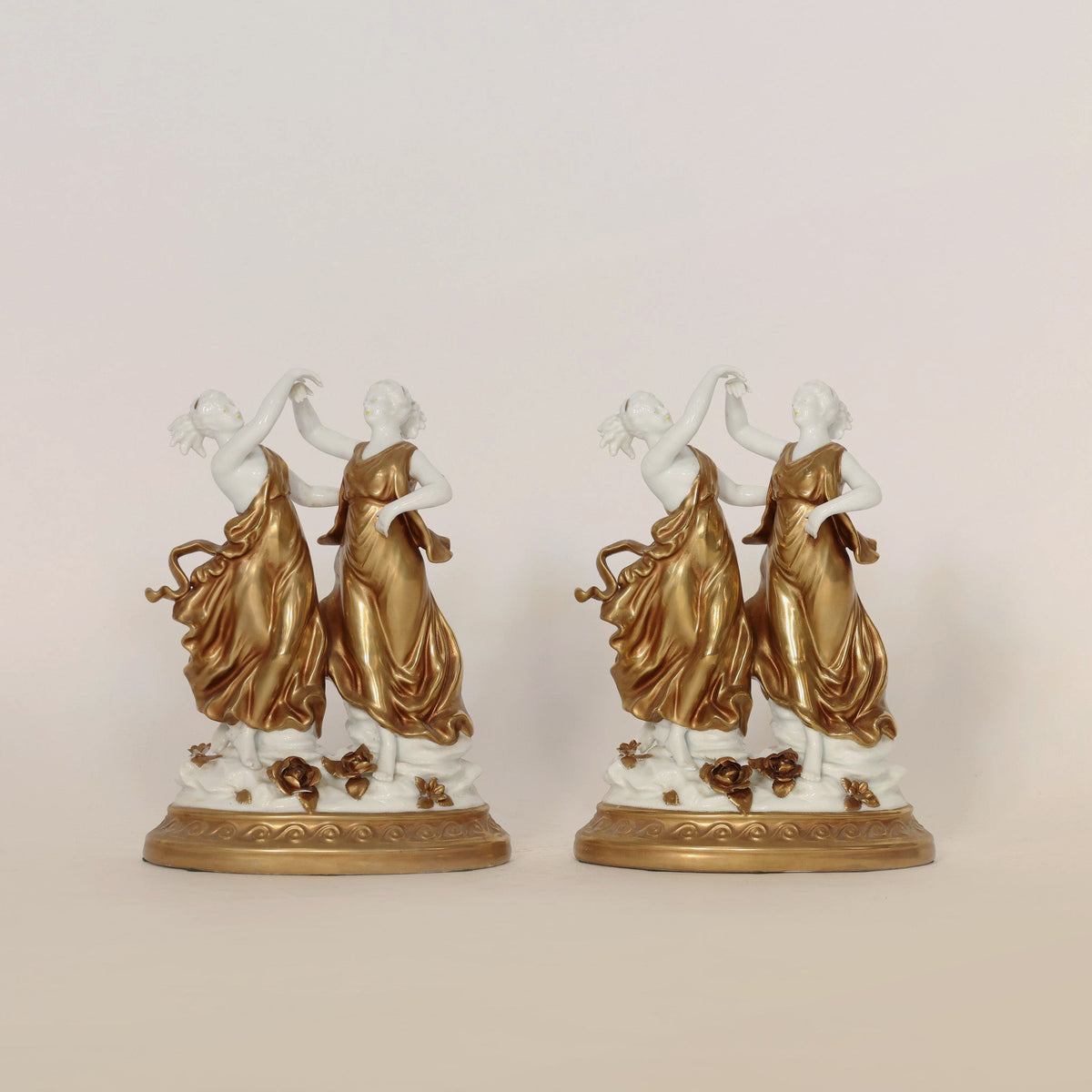 The Pair of Gilt & Porcelain Dancing Figures by Euro Trash Annie features two identical porcelain women in flowing gold and white dresses atop ornate round bases, each adorned with refined accents and gilt detailing. One woman raises her hand gracefully toward the other, capturing an elegant moment in motion.