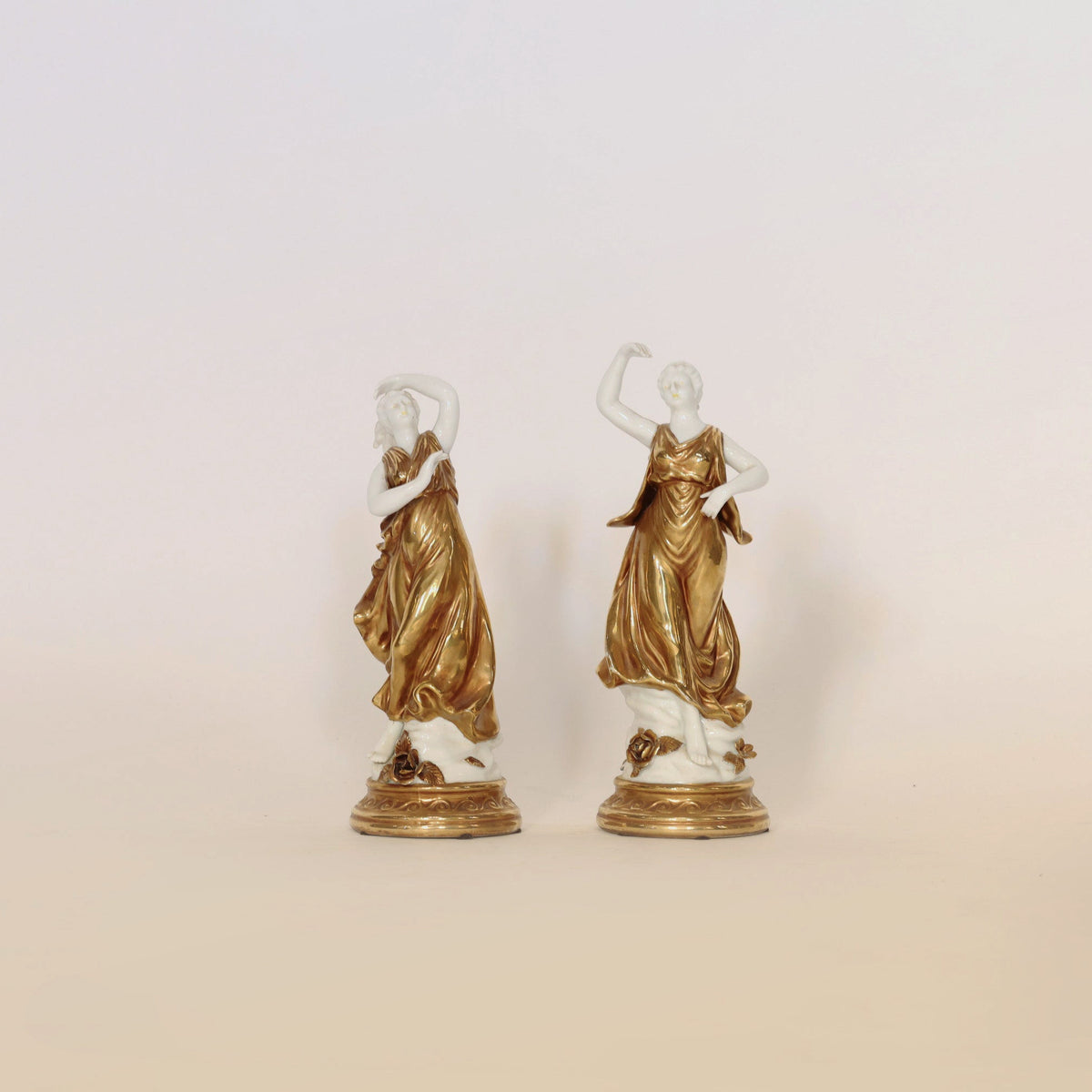 The Pair of Gilt & Porcelain Figural Sculptures by Euro Trash Annie features two elegant women in flowing gold dresses, their exquisite gilt detailing and matching ornate bases making them striking decorative accents against a plain light background.