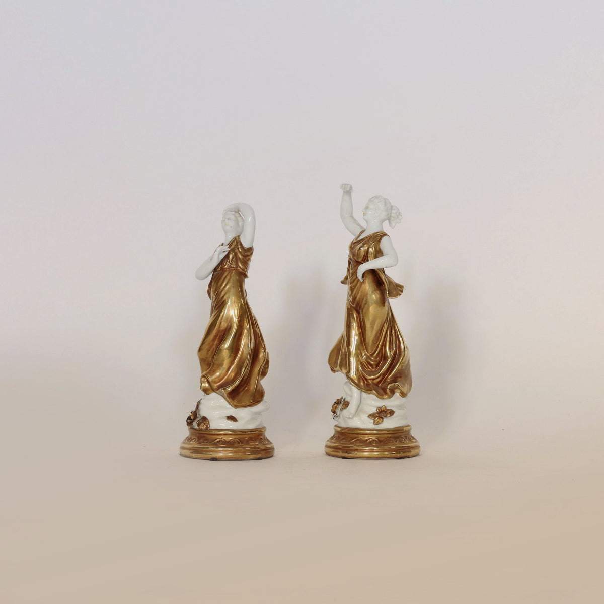 The Pair of Gilt & Porcelain Figural Sculptures by Euro Trash Annie features two women in flowing gold dresses, each gracefully posed with one arm raised. Set atop ornate round bases, these timeless pieces are enhanced by elegant gilt detailing and displayed against a plain off-white background.