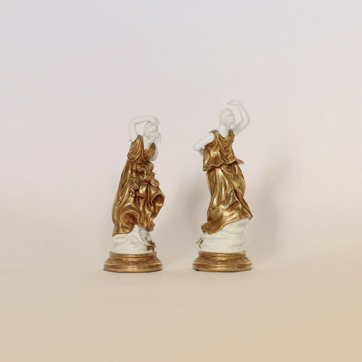 The Pair of Gilt & Porcelain Figural Sculptures by Euro Trash Annie showcases two women in flowing dresses, each standing gracefully on round bases. Enhanced with elegant gilt detailing, both figures are delicately posed with one arm raised against a simple, neutral backdrop.