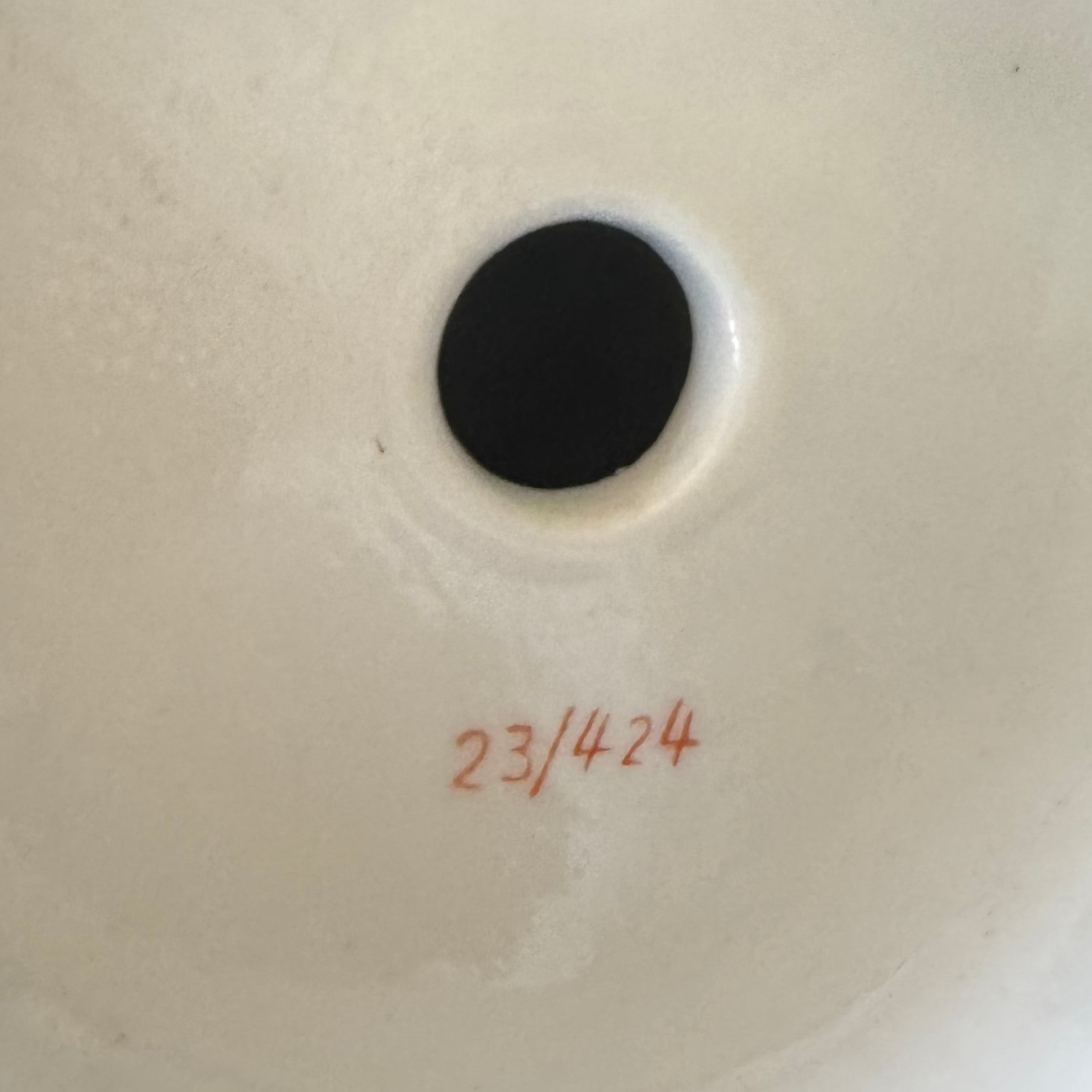 A close-up of a white surface from Euro Trash Annie's Pair of Gilt & Porcelain Figural Sculptures, showing a black circular hole near the center and "23/424" handwritten in red below, evoking subtle decorative details.