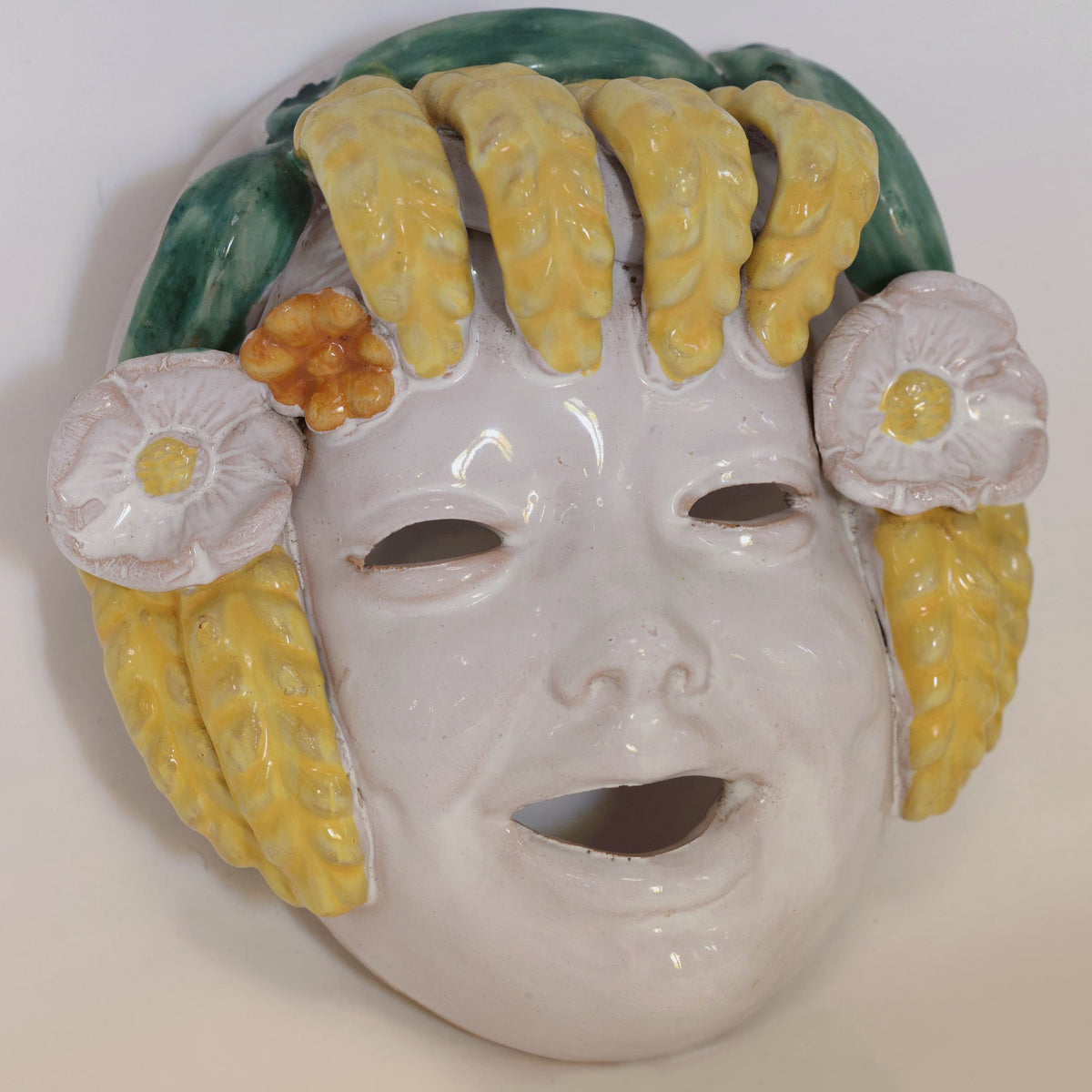 A pair of Italian Majolica figural wall masks by Euro Trash Annie, each featuring ceramic smiling faces with cut-out eyes and mouths, are adorned with vibrant yellow, green, and orange elements that resemble hair and flowers. Reminiscent of traditional Italian majolica craftsmanship, these striking pieces of artisanal wall art infuse any space with vivid color and playful design, celebrating joyful artistry in home decor.