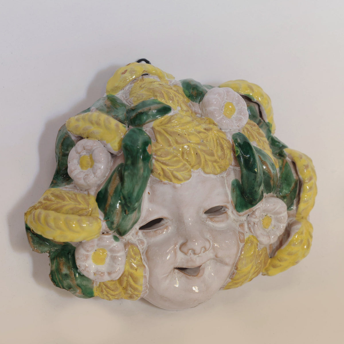 A pair of Italian Majolica figural wall masks by Euro Trash Annie, each depicting a child’s face adorned with yellow and green leaves and white flowers for hair, features expressive open mouths and hollow eyes. Hung against a simple backdrop, these artisanal pieces bring the timeless charm of Italian craftsmanship to your home decor.