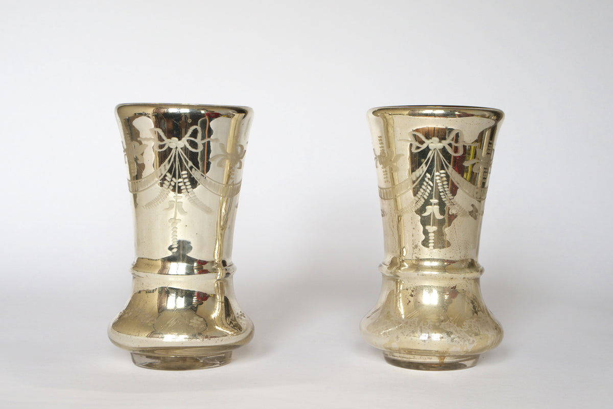 The Pair of Vintage Etched Mercury Glass Vases by Euro Trash showcases a shiny silvered finish, ornate bows, and a garland motif, elegantly displayed against a plain white background.