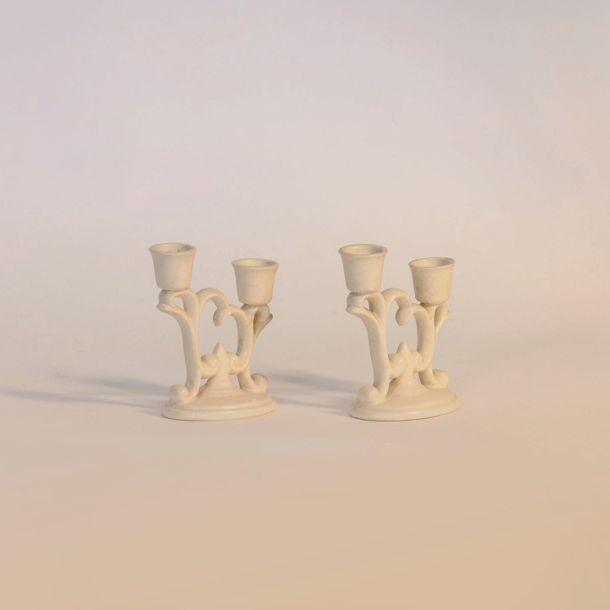 A Pair of Vintage Ivory-Tone Candelabras by Euro Trash Annie, featuring ornate, curved designs and each holding two taper candles, are displayed on a neutral background. These elegant candelabras add timeless charm as sophisticated candle holders to any space.