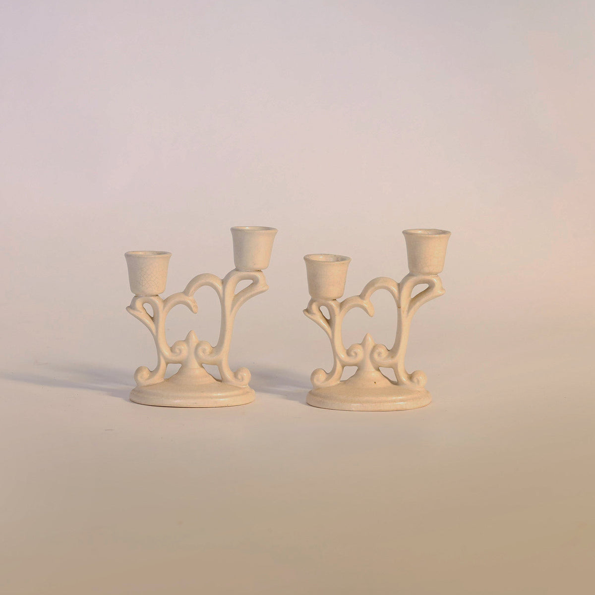 A pair of Euro Trash Annie Vintage Ivory-Tone Candelabras, each featuring two arms and an ornate ceramic design, are displayed side by side on a light surface with a subtle gradient background.