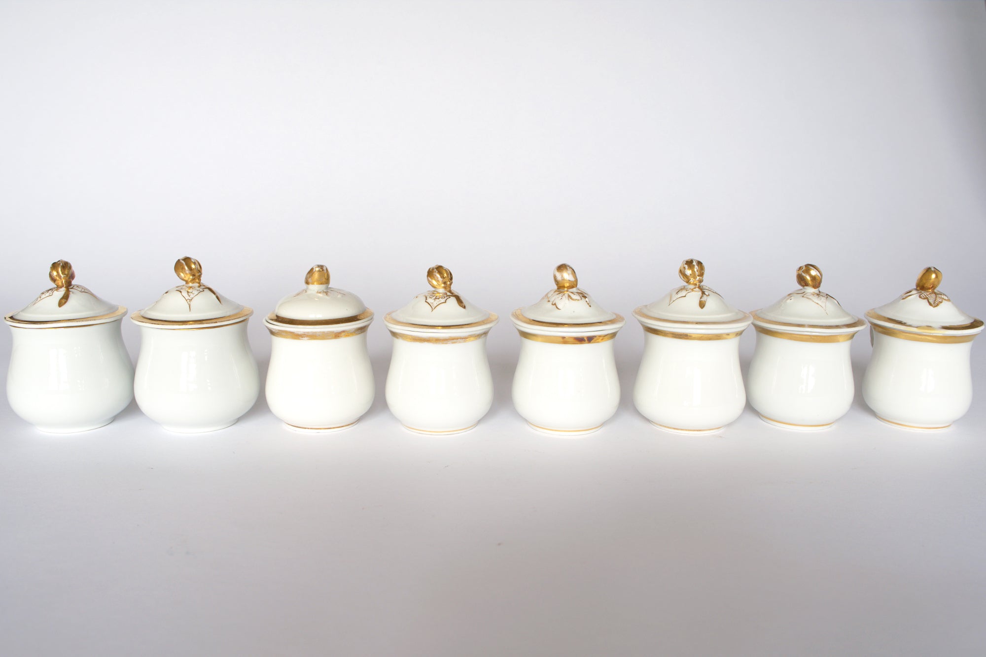 The Set of Breakfast Pots by Euro Trash features eight white porcelain pots with lids, each accented by a gold finial, arranged in a row against a plain white background.
