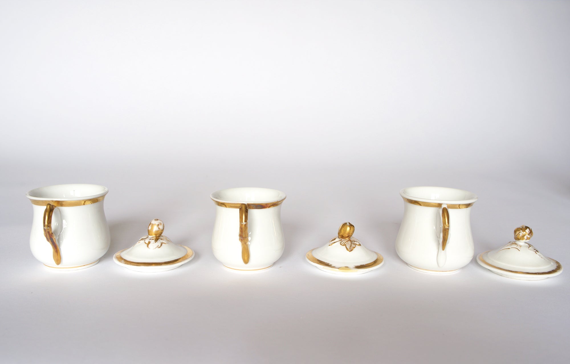 The Euro Trash Set of Breakfast Pots features three white porcelain pots with gold accents, arranged in a row. Two lids are placed in front, and the rightmost pot has its lid tilted to the side, all set against a white background.