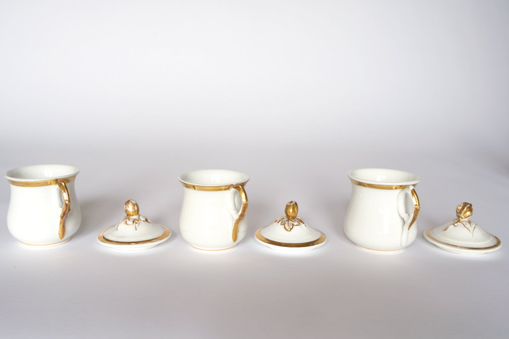 The Set of Breakfast Pots by Euro Trash features four white porcelain pots with gold accents and beetle-shaped finial lids, arranged in a row against a white background, with some lids on the pots and others placed beside them.
