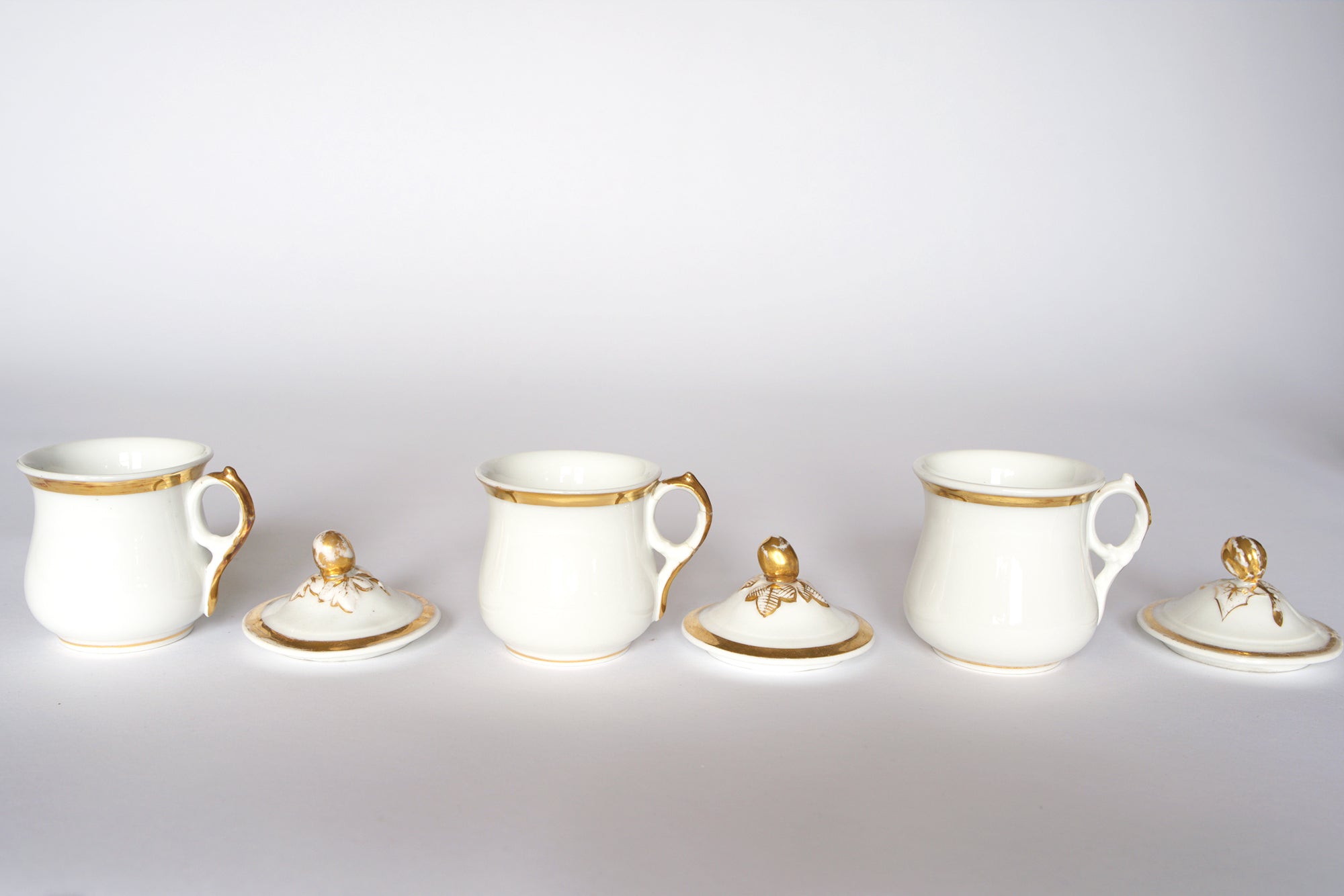 The Euro Trash Set of Breakfast Pots features three elegant white porcelain pots with gold rims, matching lids, and gold finials, beautifully arranged in a row against a plain white background.