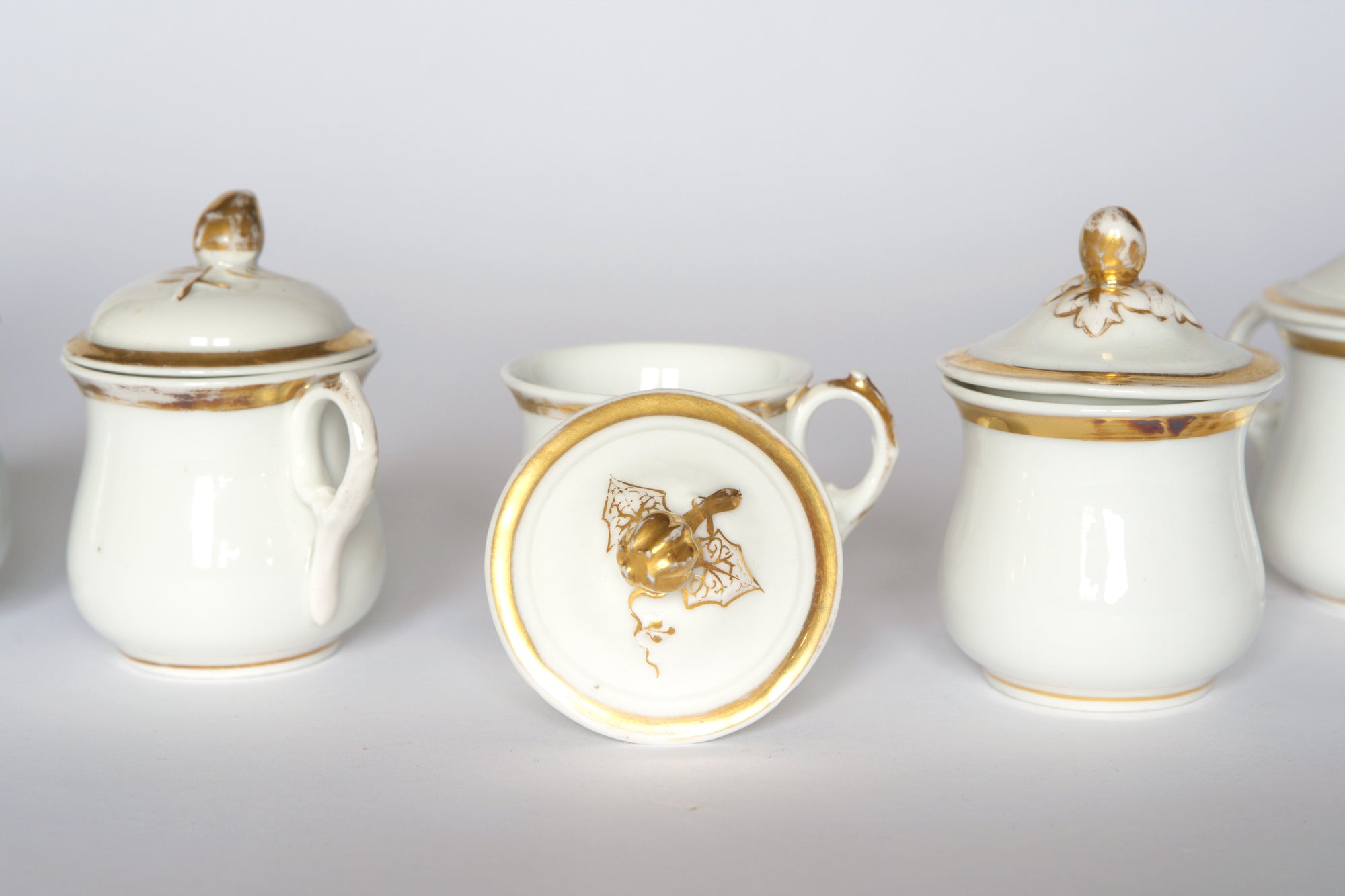 The Set of Breakfast Pots by Euro Trash features white porcelain pots with handles and gold accents, including elegant lids—one upright to reveal a gold bat decoration—and decorative gold finials.