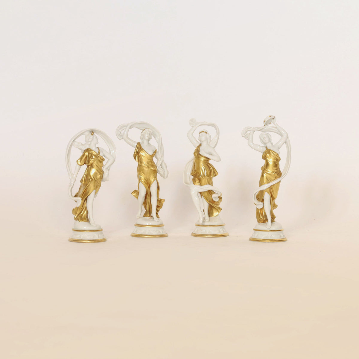 The Set of Four Gilt & Porcelain Figurines by Euro Trash Annie features four elegant sculptures of women, each posed uniquely in flowing gold and white drapery. The exquisite gilt detailing enhances their charm, making them stunning decorative collectibles against a simple light background.