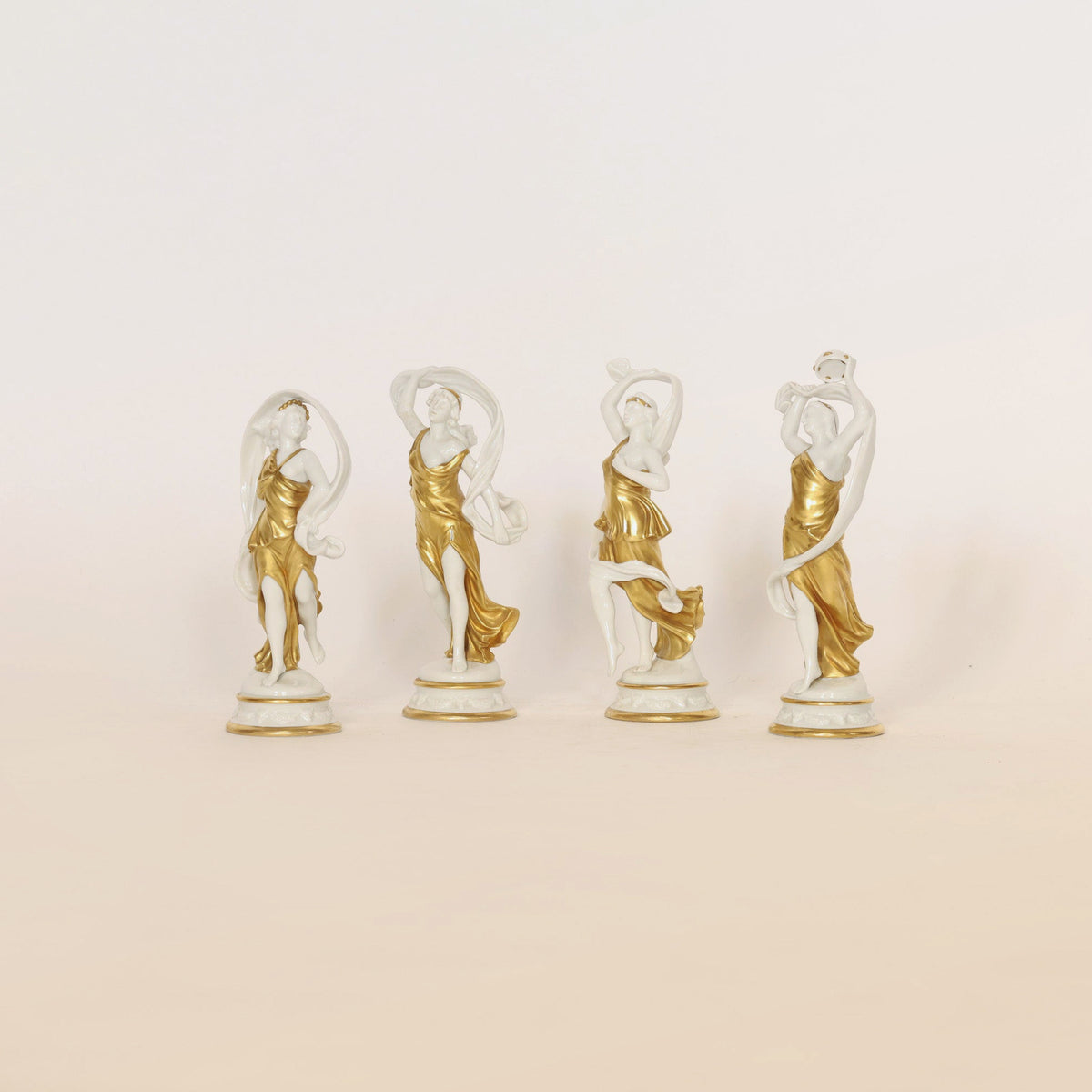 A set of four elegant gilt and porcelain figurines by Euro Trash Annie features women in gold dresses, each adorned with fine gilt detailing. These decorative collectibles are gracefully posed in a row against a simple, light background, each figure holding up fabric above her head.