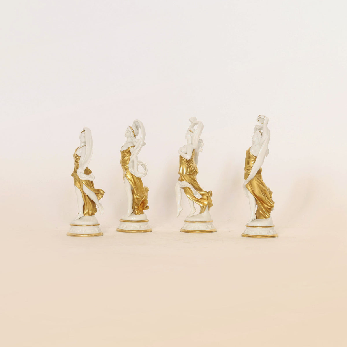 Four neoclassical-style porcelain figurines from Euro Trash Annie’s “Set of Four Gilt & Porcelain Figurines” are displayed in a row against a plain background. Each decorative piece features an elegantly posed female figure in a flowing white and gold gown with intricate gilt detailing, poised gracefully atop a gold-rimmed pedestal.