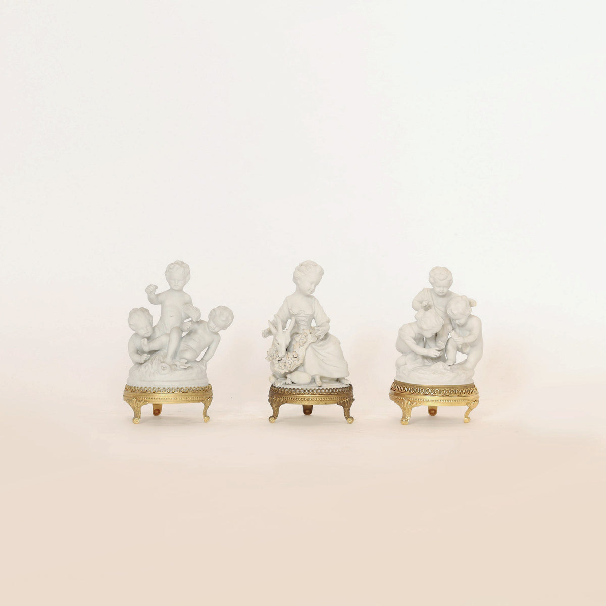 The Set of Porcelain Cherub Figurines on Ormolu Bases by Euro Trash Annie features three white porcelain figurines, each with gold-trimmed ormolu bases and depicting multiple cherubic children in playful or gentle poses. Displayed against a plain white background, these decorative collectibles bring an elegant touch to any setting.