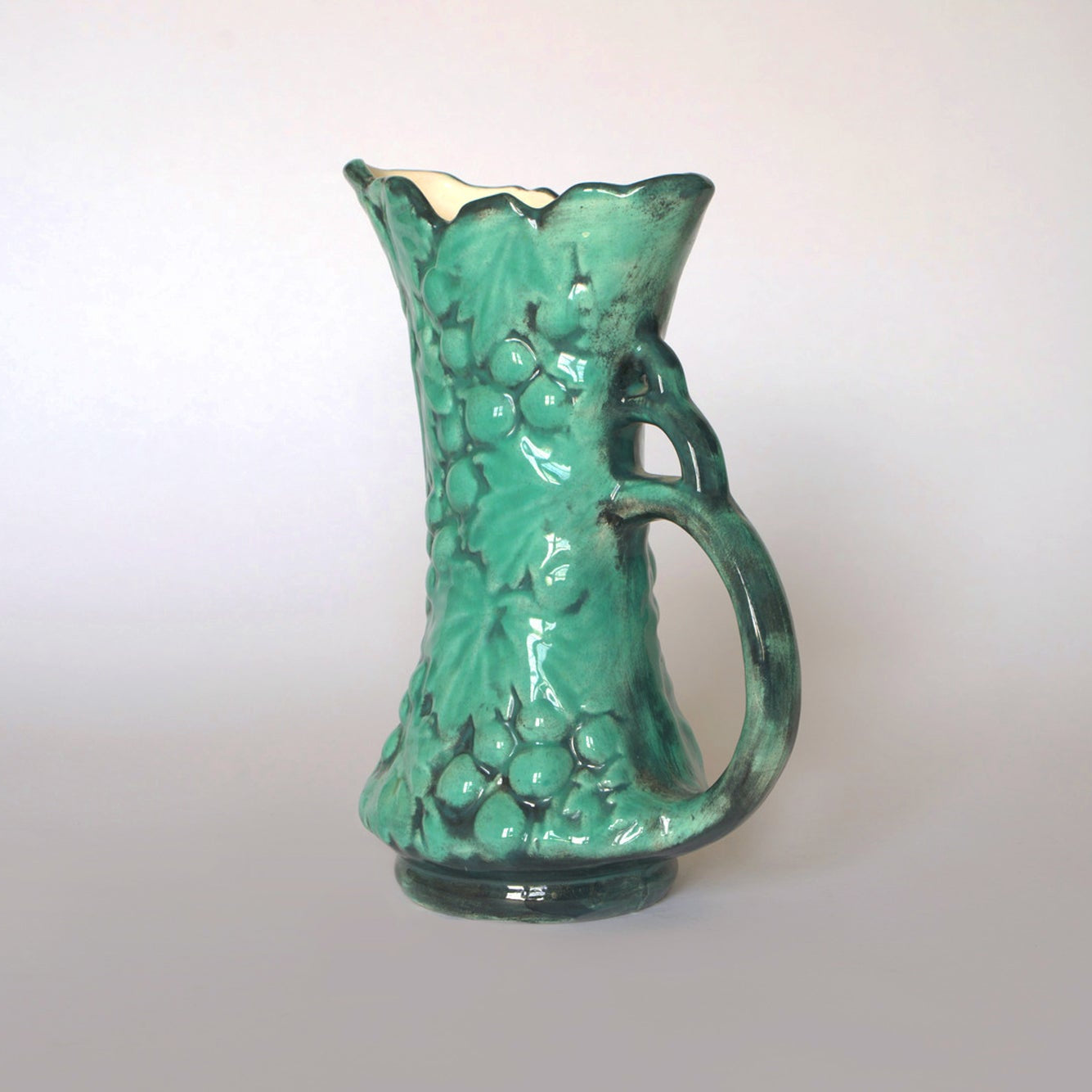 The Euro Trash Vintage Green Majolica Style Grape Motif Ceramic Pitcher features a sculpted handle, raised grape clusters, and leaves for a classic look, displayed against a simple light background.