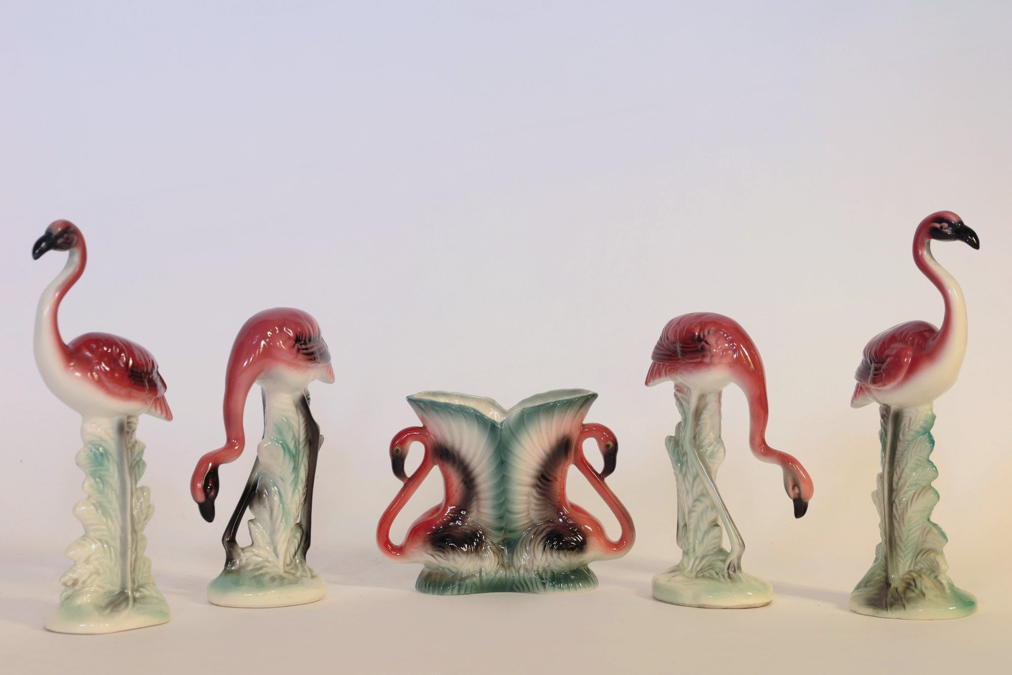 The Euro Trash Annie Vintage Flamingo Figurines & Vase Set features five charming flamingo figurines arranged in a row with a pink and green leaf-shaped decorative vase at the center. Each flamingo faces a different direction against a plain background, creating an eye-catching sculptural home decor display.
