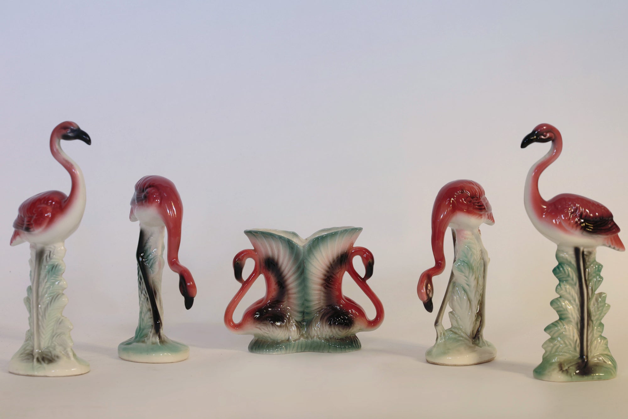 The Vintage Flamingo Figurines & Vase Set by Euro Trash Annie features five ceramic flamingo pieces arranged in a row: three stand upright, two are bent down, and the central vase showcases a pair of flamingos with their necks gracefully curved inward. This sculptural set infuses any space with nostalgic charm and playful elegance.