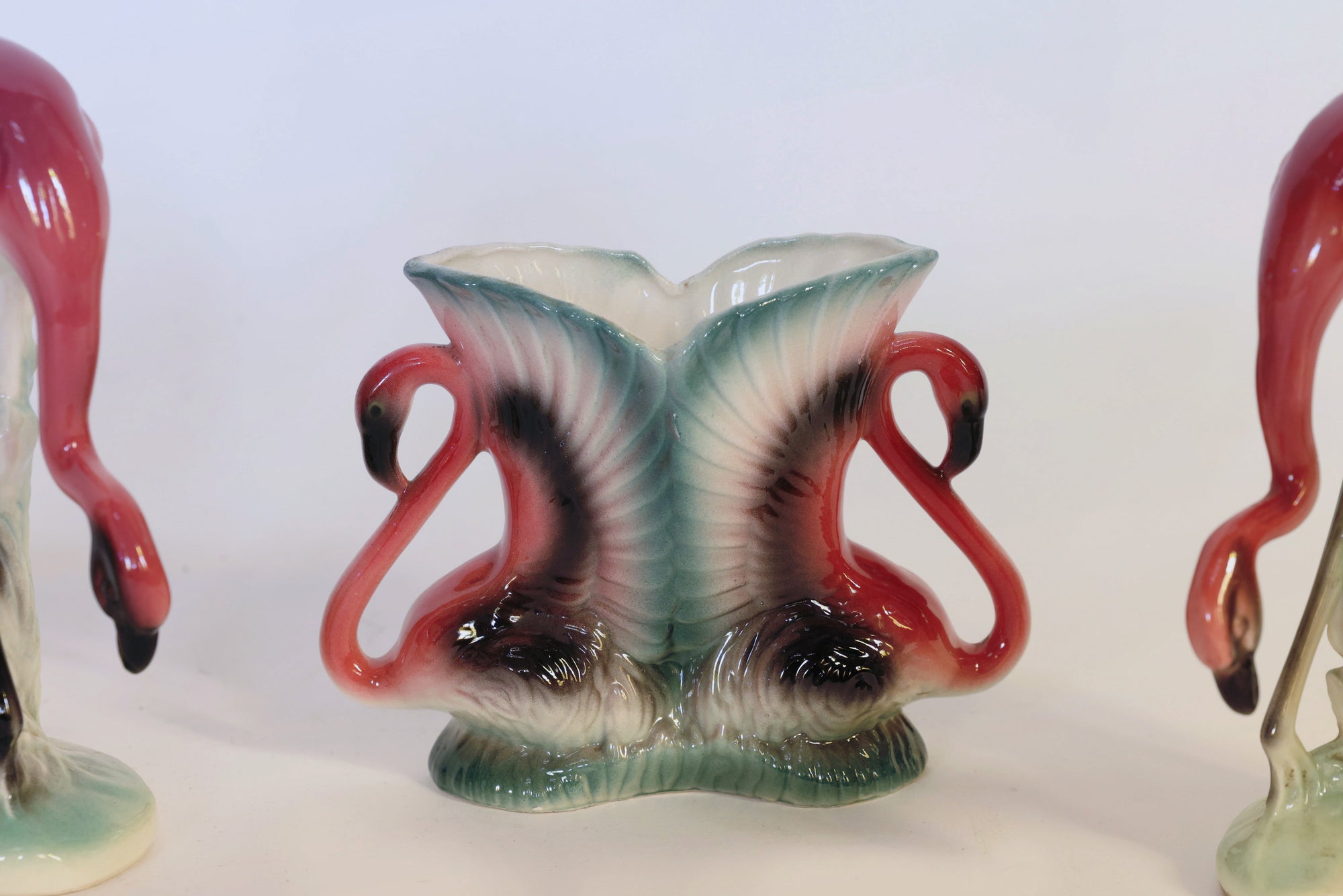 The Vintage Flamingo Figurines & Vase Set by Euro Trash Annie features a ceramic vase shaped like two pink flamingos with their necks elegantly curved into a heart, accompanied by matching vintage flamingo figurines on either side, creating a charming sculptural home decor display.