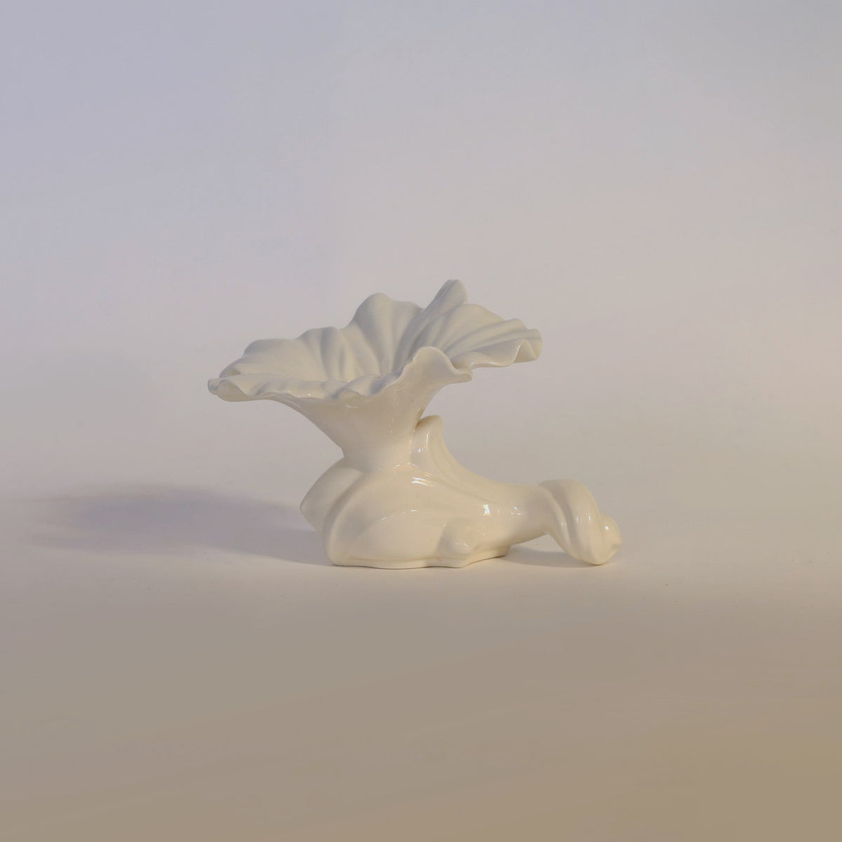 The Euro Trash Annie Vintage Milk Glass Ruffled Vase features a glossy white ceramic finish with a ruffled edge, echoing the delicate form of a flower unfurling from an abstract, curved base. Set against a plain light background, its opaque surface exudes the timeless elegance of classic milk glass design.