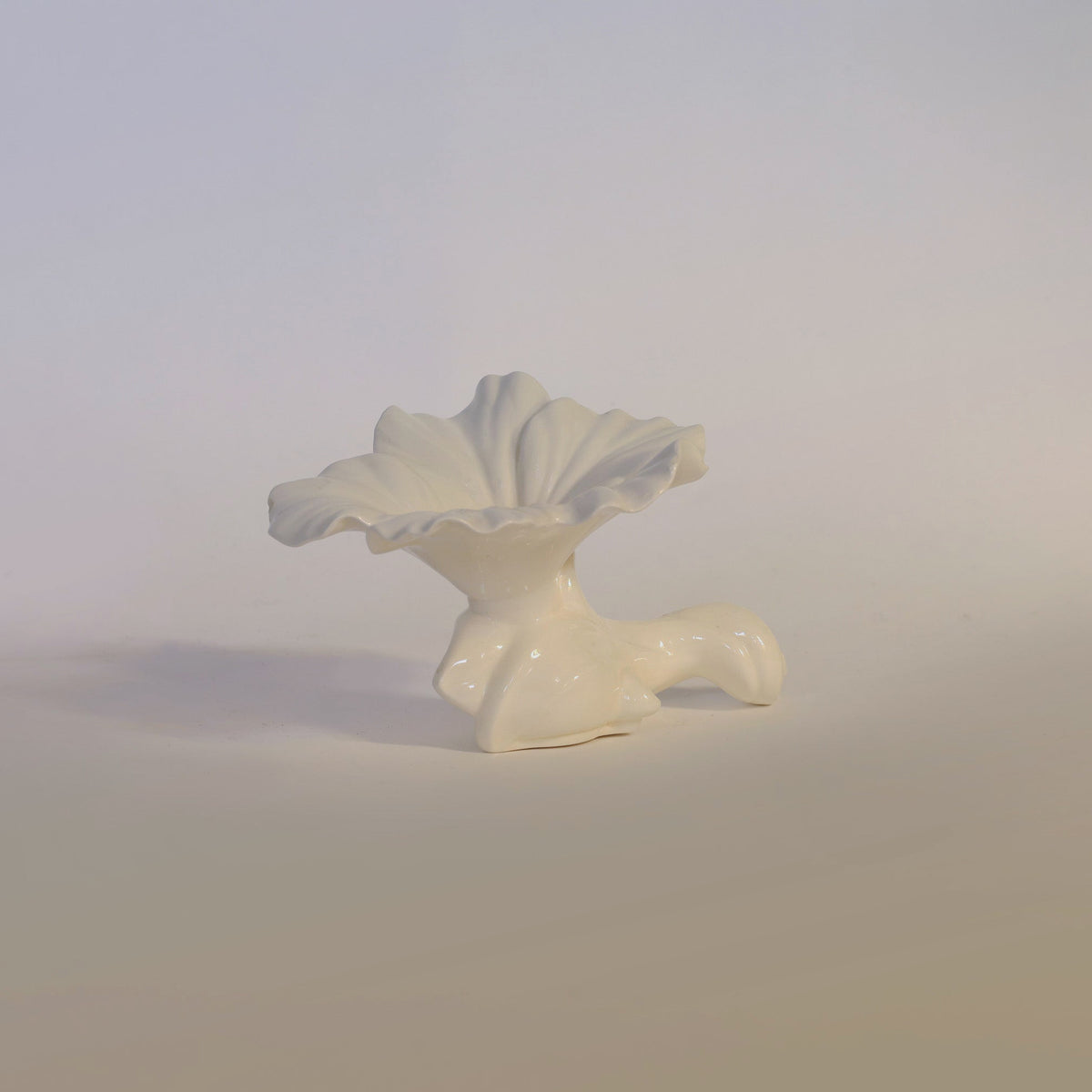 A sculpted white Vintage Milk Glass Ruffled Vase by Euro Trash Annie, featuring petal-like ruffled edges and a curved, stem-inspired base, is set against a plain light background. Its opaque finish exudes the timeless charm associated with classic milk glass.