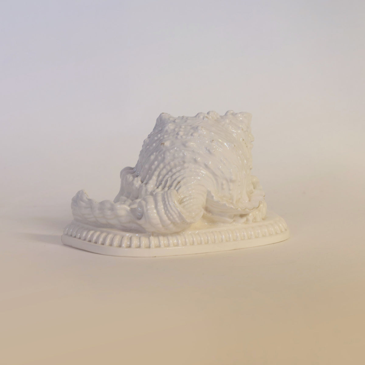 The Euro Trash Annie Vintage Milk Glass Seashell Sculpture features a glossy white ceramic conch shell design, reminiscent of classic milk glass seashell decor, elegantly displayed on a matching oval base against a light beige background.