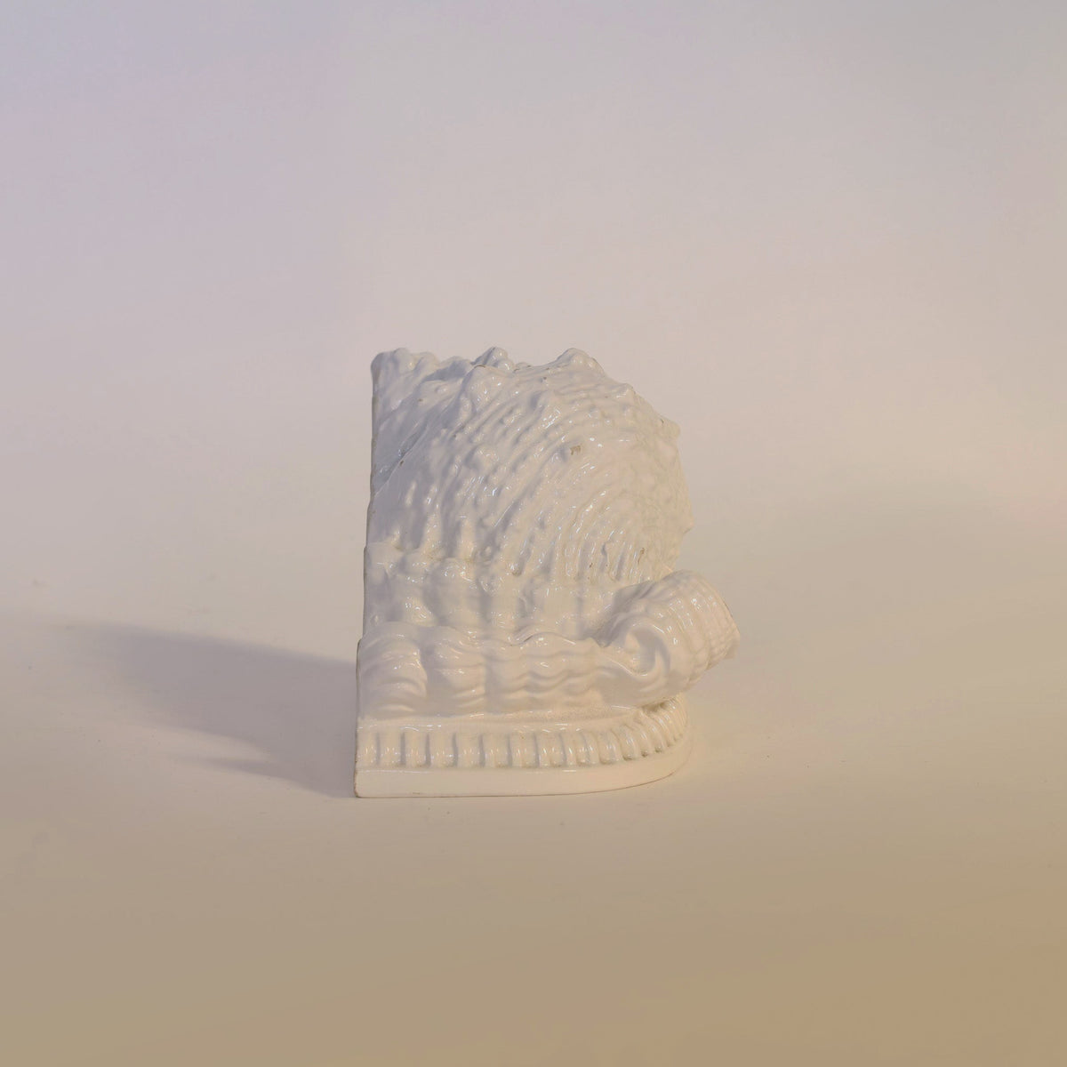 The Vintage Milk Glass Seashell Sculpture by Euro Trash Annie features a white ceramic wedge shape with textured detailing and a rounded, coiled accent reminiscent of vintage milk glass seashells, making it an ideal sculptural accent for coastal decor. Displayed on a light, neutral background.