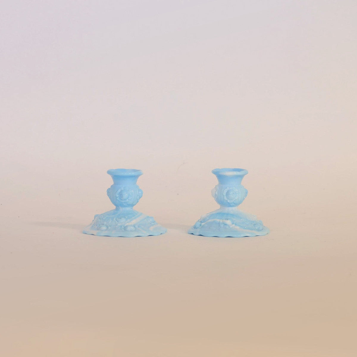 A pair of vintage blue milk glass candlestick holders by Euro Trash Annie, featuring ornate bases, are positioned side by side on a neutral beige surface against a plain pale background.