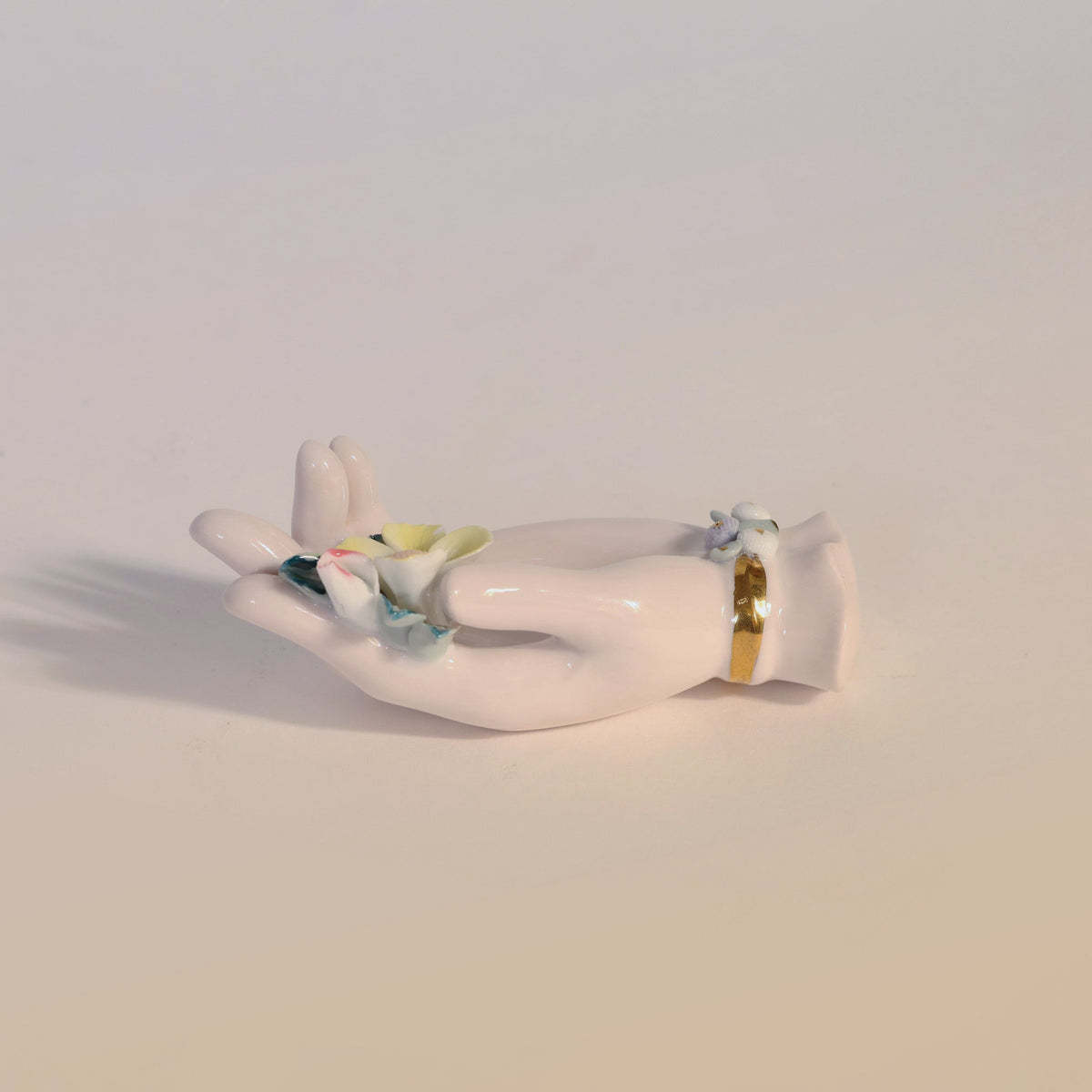 The Vintage Porcelain Hand Ring Holder by Euro Trash Annie features a delicate pastel flower and a gold bracelet detail, resting elegantly on a light surface and casting a soft shadow—a charming decorative accent for any space.